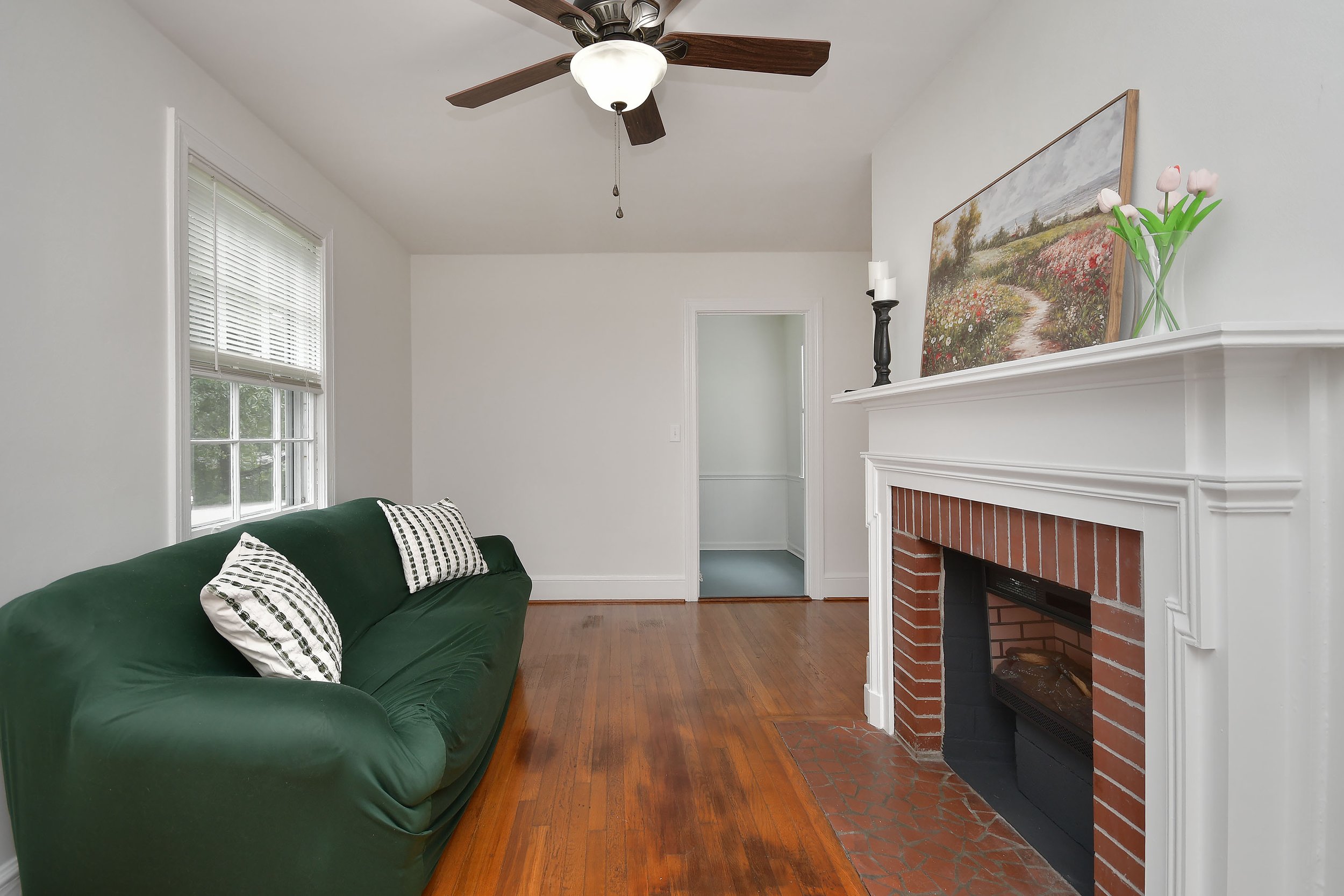 A cozy living room with a green sofa with black and white pillows, a white fireplace with a painting on top, a vase of pink tulips, a black candle holder, a ceiling fan, a window with blinds, and hardwood flooring.