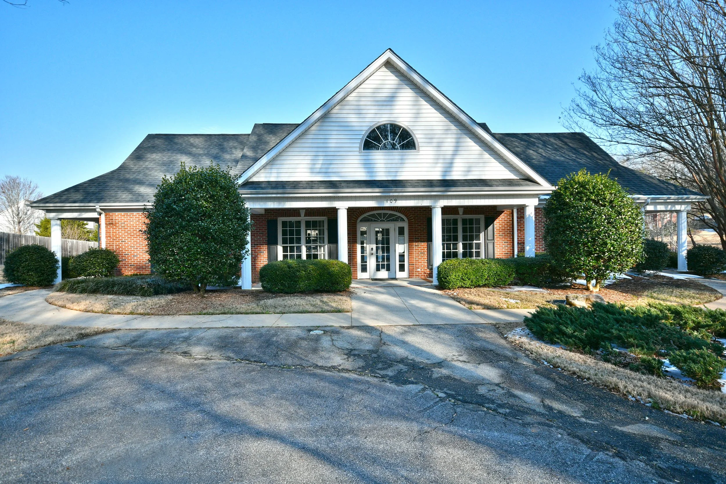 Front view of a house with a gabled roof, white siding and brick facade, surrounded by landscaped bushes and trees, with a cracked asphalt driveway in the foreground. Home for sale in Greenville, SC. Master on Main Level. 4 Bedroom home. Upscale.