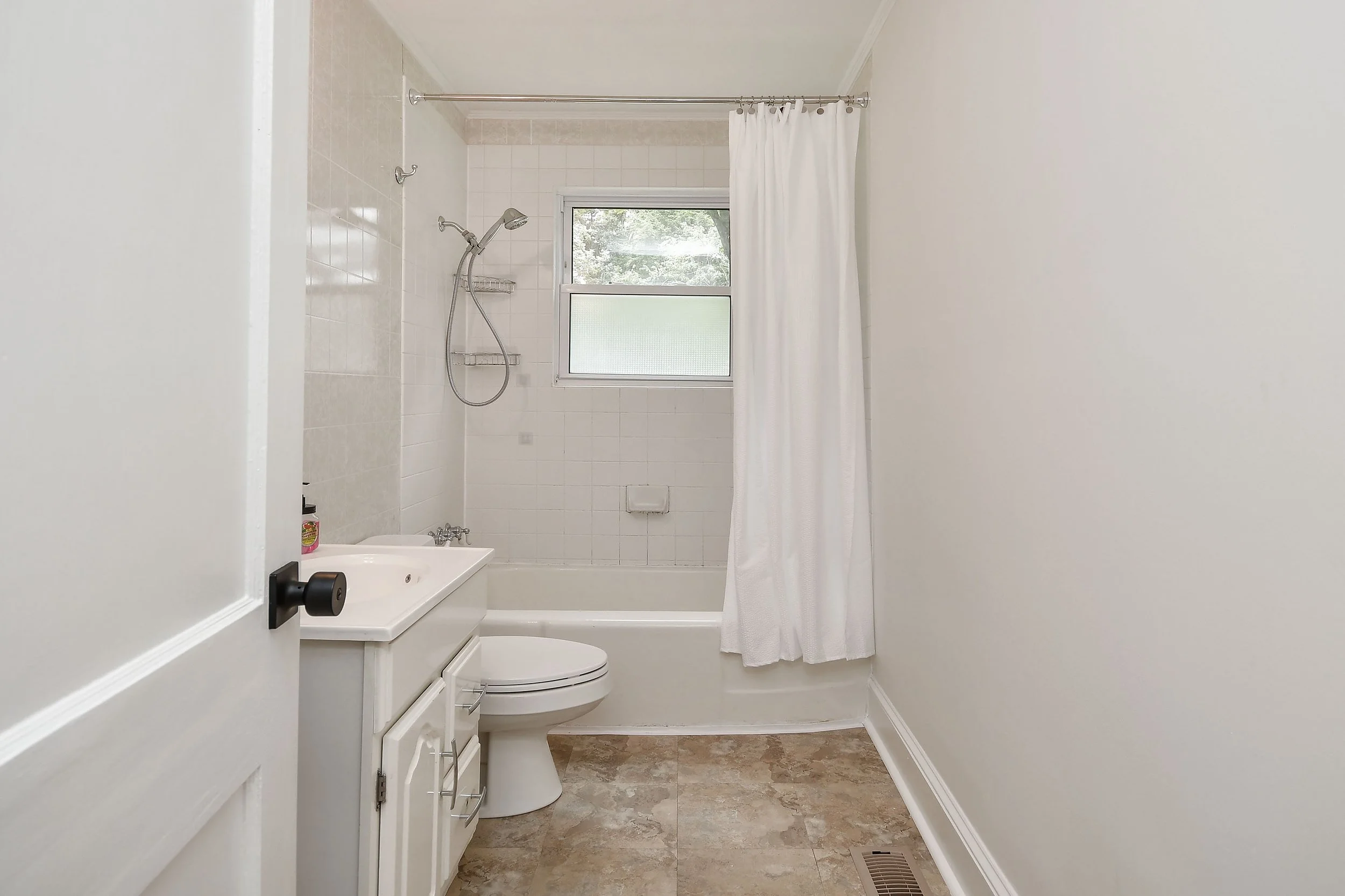A small bathroom with a white bathtub, a shower head, and a white shower curtain. There is a window with frosted glass and a view of trees outside. The bathroom has beige tiled flooring and white walls.