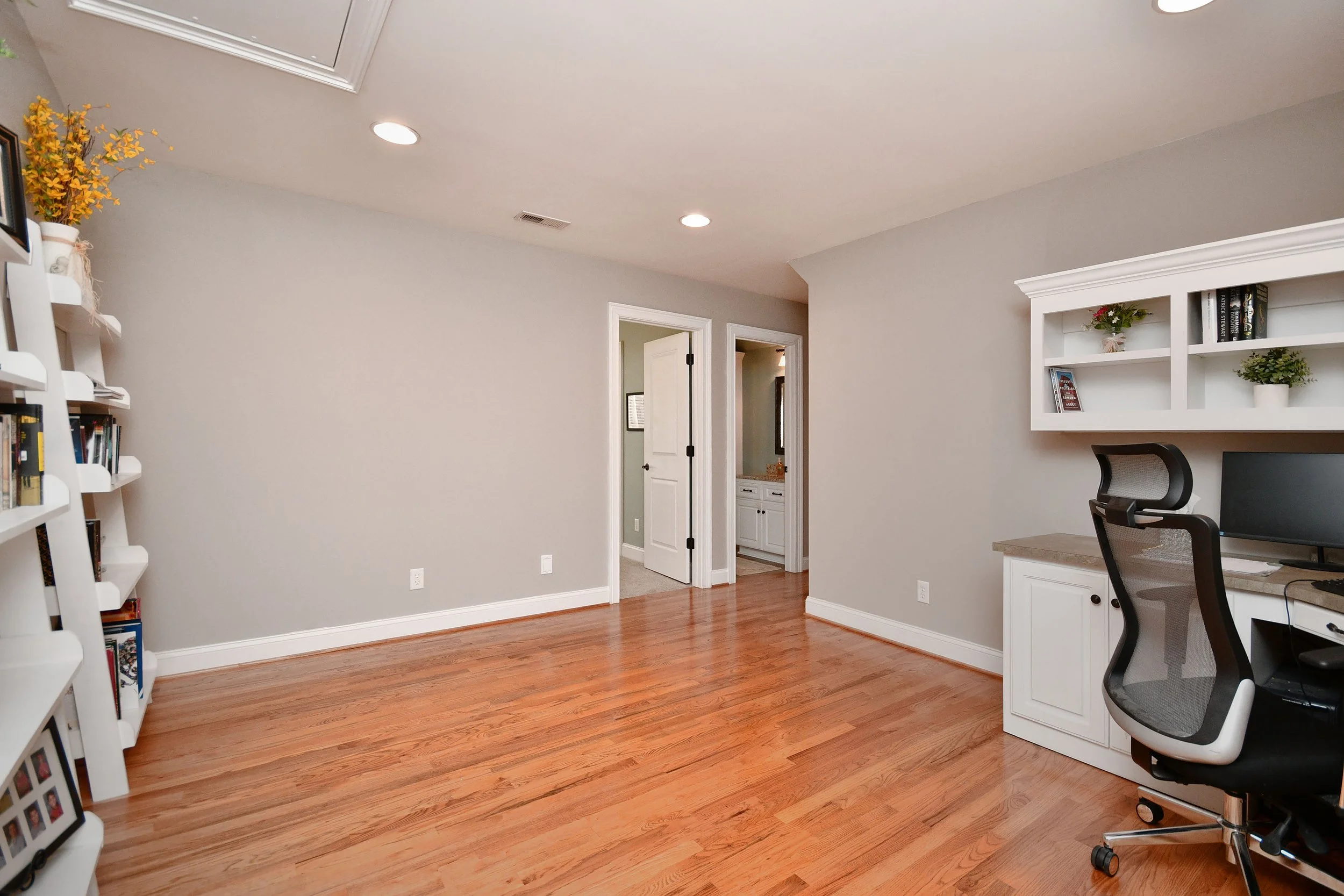 Empty room with hardwood floor, gray walls, a white desk with a computer and an ergonomic chair, a white wall shelf with decorative items, a doorway leading to a bathroom, recessed lighting, and a bookshelf with framed photos and plants. 4 Bed