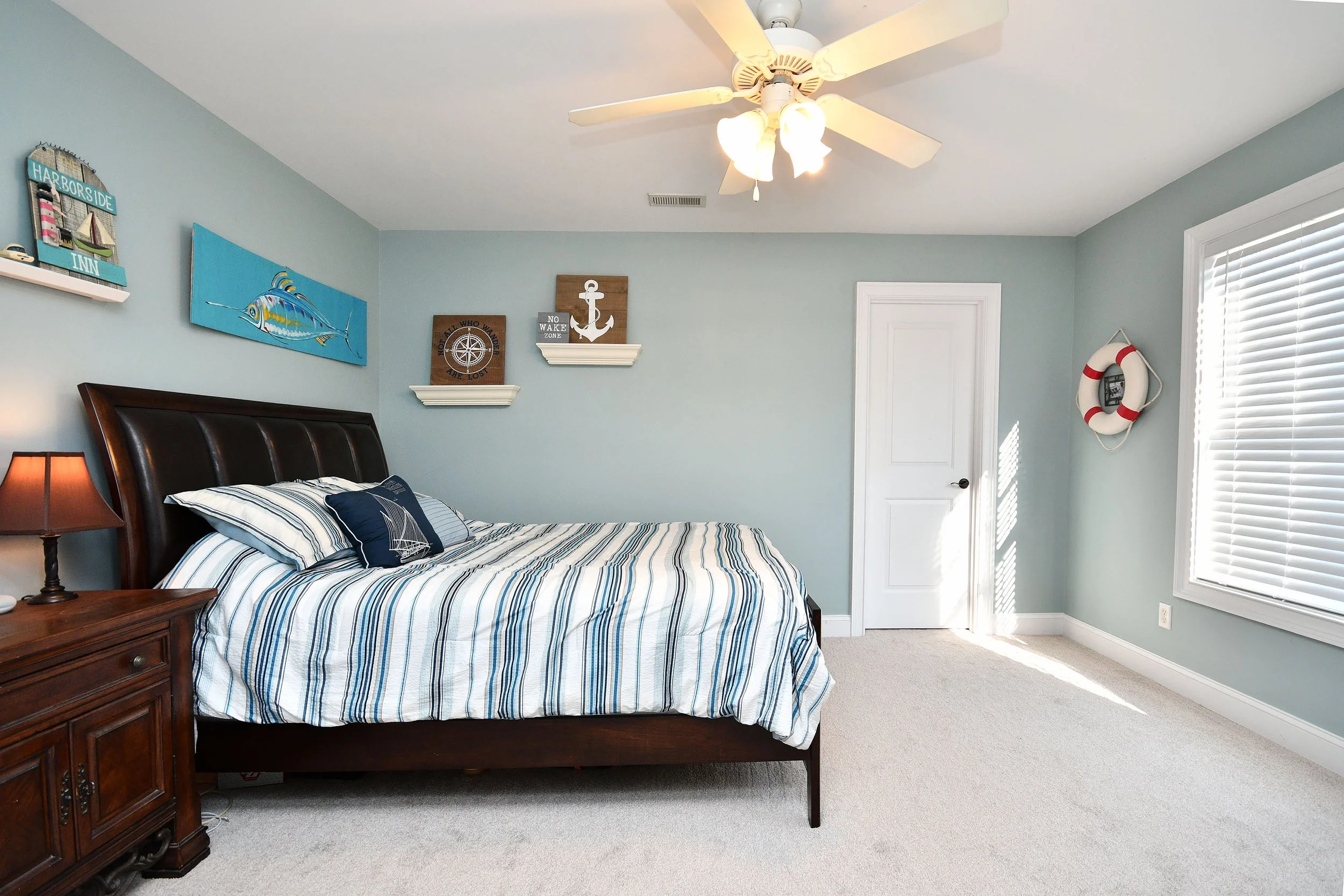 A bedroom with a wooden bed, striped bedding, a nightstand with a lamp, wall decorations with a nautical theme, and a ceiling fan; bright sunlight coming through the window. Home for sale in Greenville, SC. Master on Main Level. 4 Bedroom home