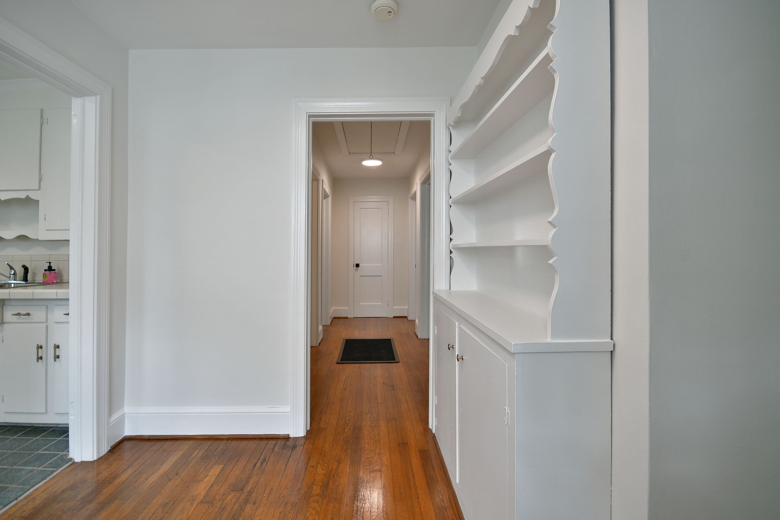 View of an interior hallway with hardwood floors, white walls, and a white built-in shelf on the right. The hallway leads to a closed door at the end and has a small black rug on the floor. Part of a kitchen with white cabinets and a sink is visible on the left.