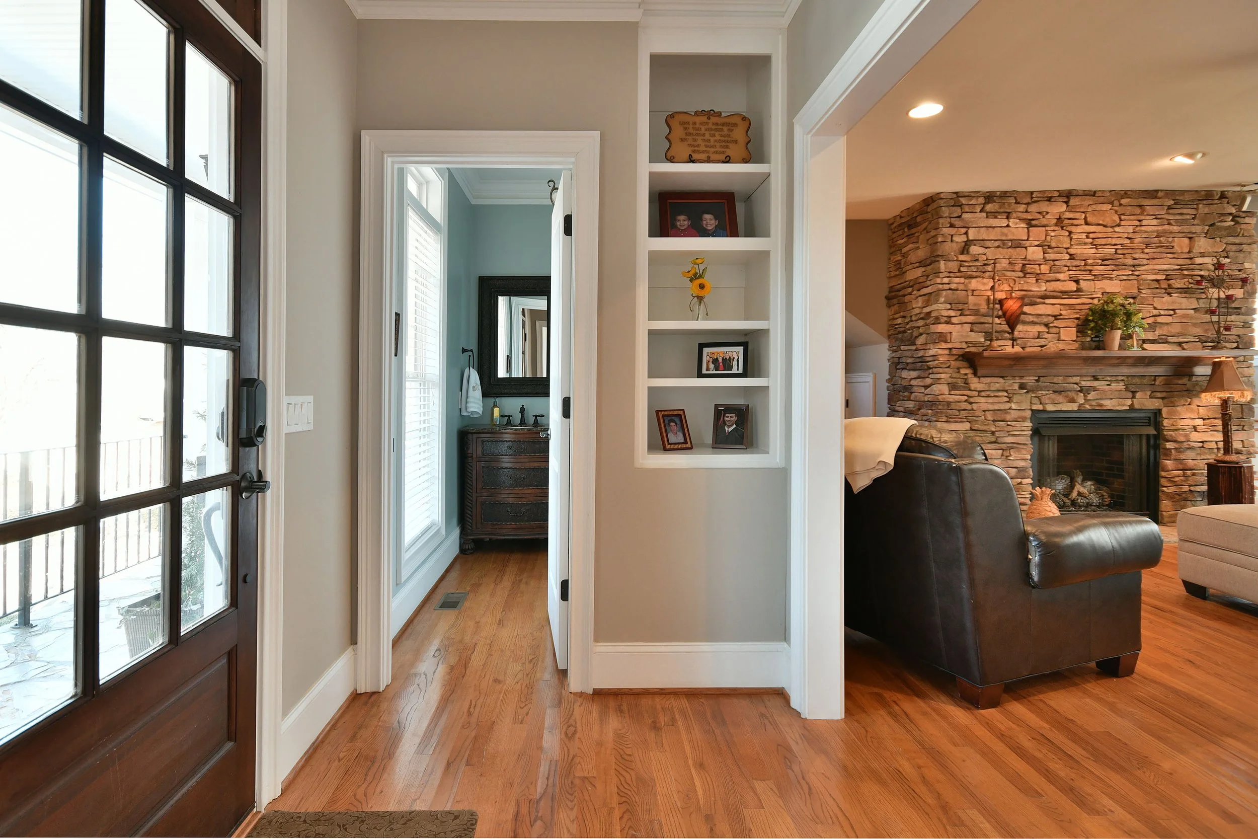 Living room with a brick fireplace, leather couch, and a shelf with framed photos, with a view of a bathroom through an open door. Home for sale in Greenville, SC. Master on Main Level. 4 Bedroom home for sale. Upscale Community. 
