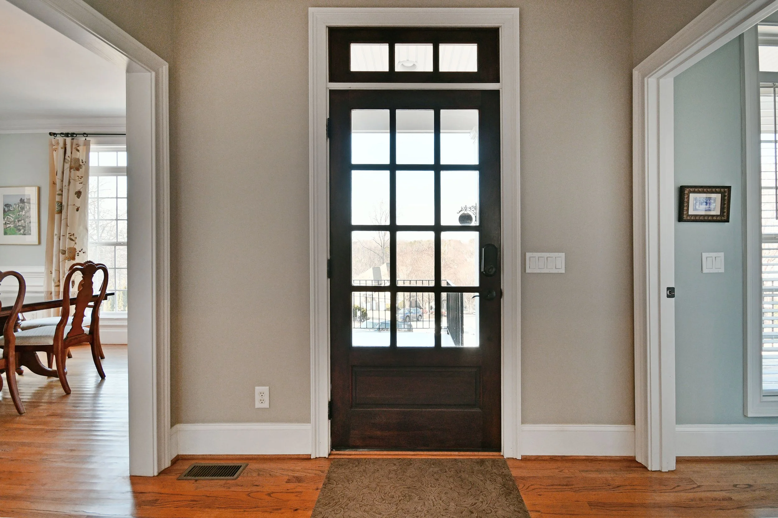 Entrance door with black frame and windowpanes, leading outside to a snowy landscape, flanked by beige and light blue walls, and adjacent to a dining room with wooden chairs and a large window. Home for sale in Greenville, SC. Master on Main Level. 