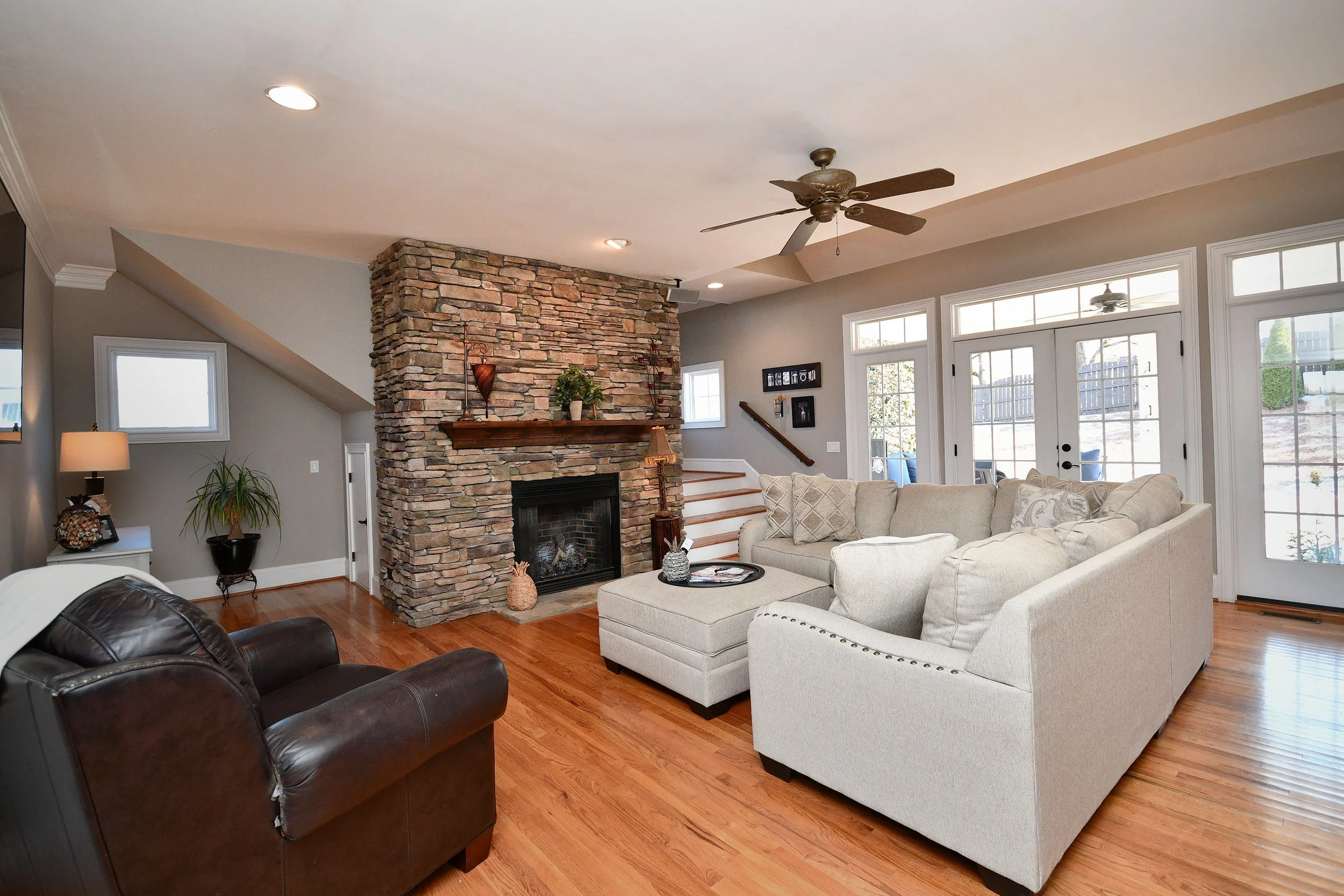 Living room with stone fireplace, beige sectional sofa, dark leather armchair, hardwood floors, ceiling fan, large glass doors, and windows. Home for sale in Greenville, SC. Master on Main Level. 4 Bedroom home for sale. Upscale Community. 