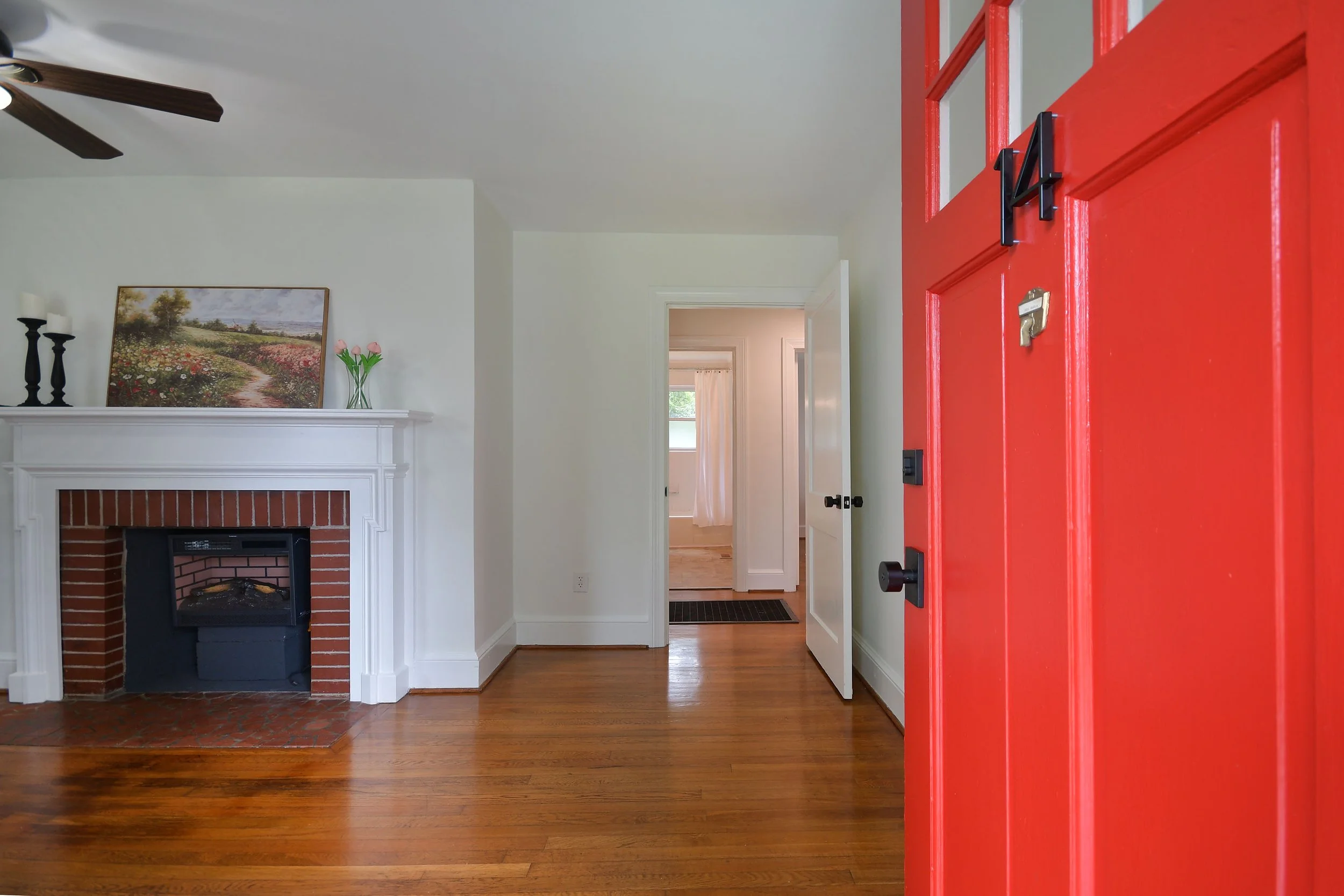 View of a living room with a red front door, hardwood floors, a white fireplace mantel with a landscape painting and candle holders, and a hallway leading to other rooms with visible windows and natural light.