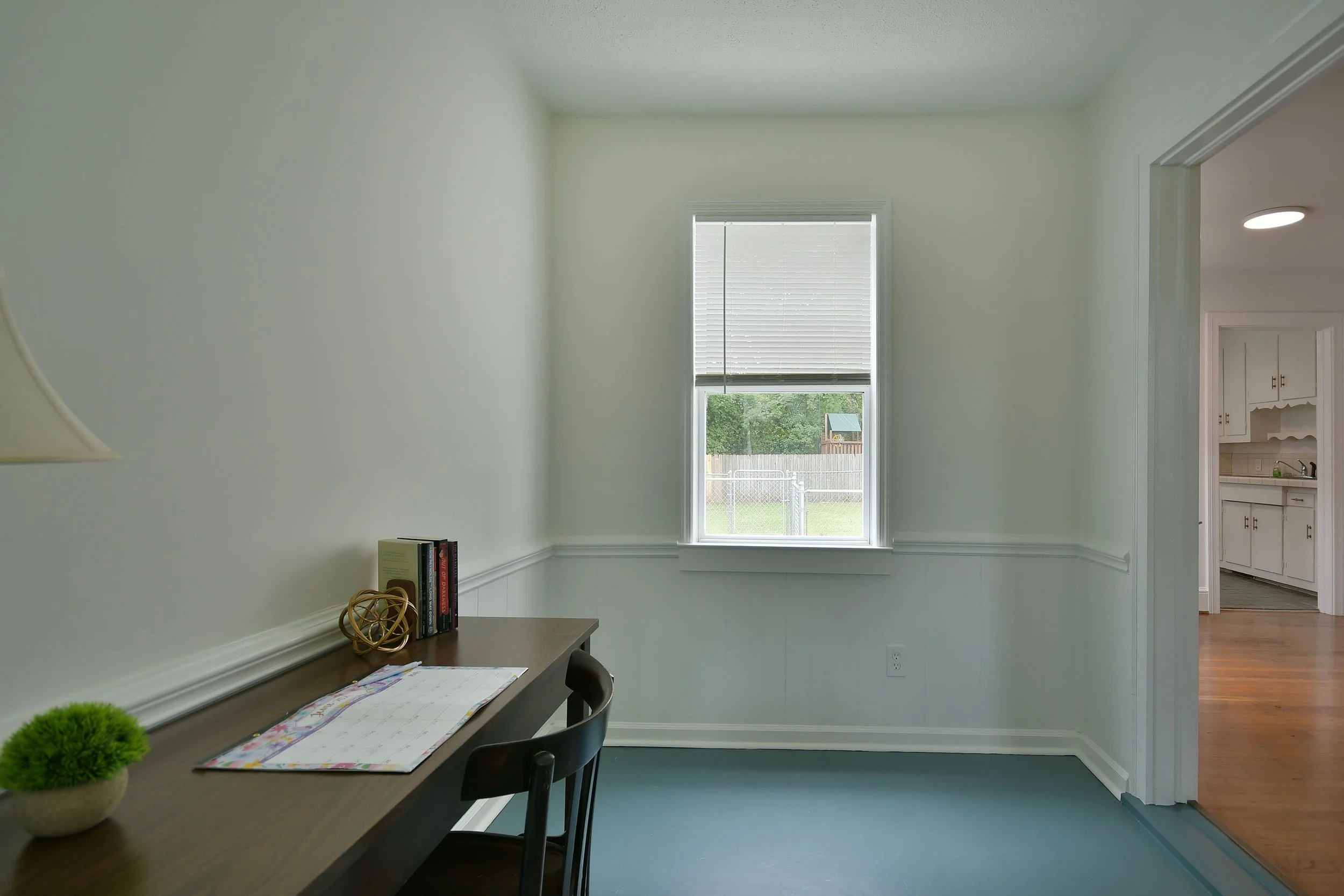 A minimalistic home office or dining area with a wooden table, two black chairs, a small potted plant, and a few books. It has a large window with blinds on the wall, letting in natural light, with a view of a fenced backyard with a small playhouse. To the right, there is part of a doorway leading to a kitchen with white cabinets and a sink. The room has white walls, crown molding, and hardwood floors.