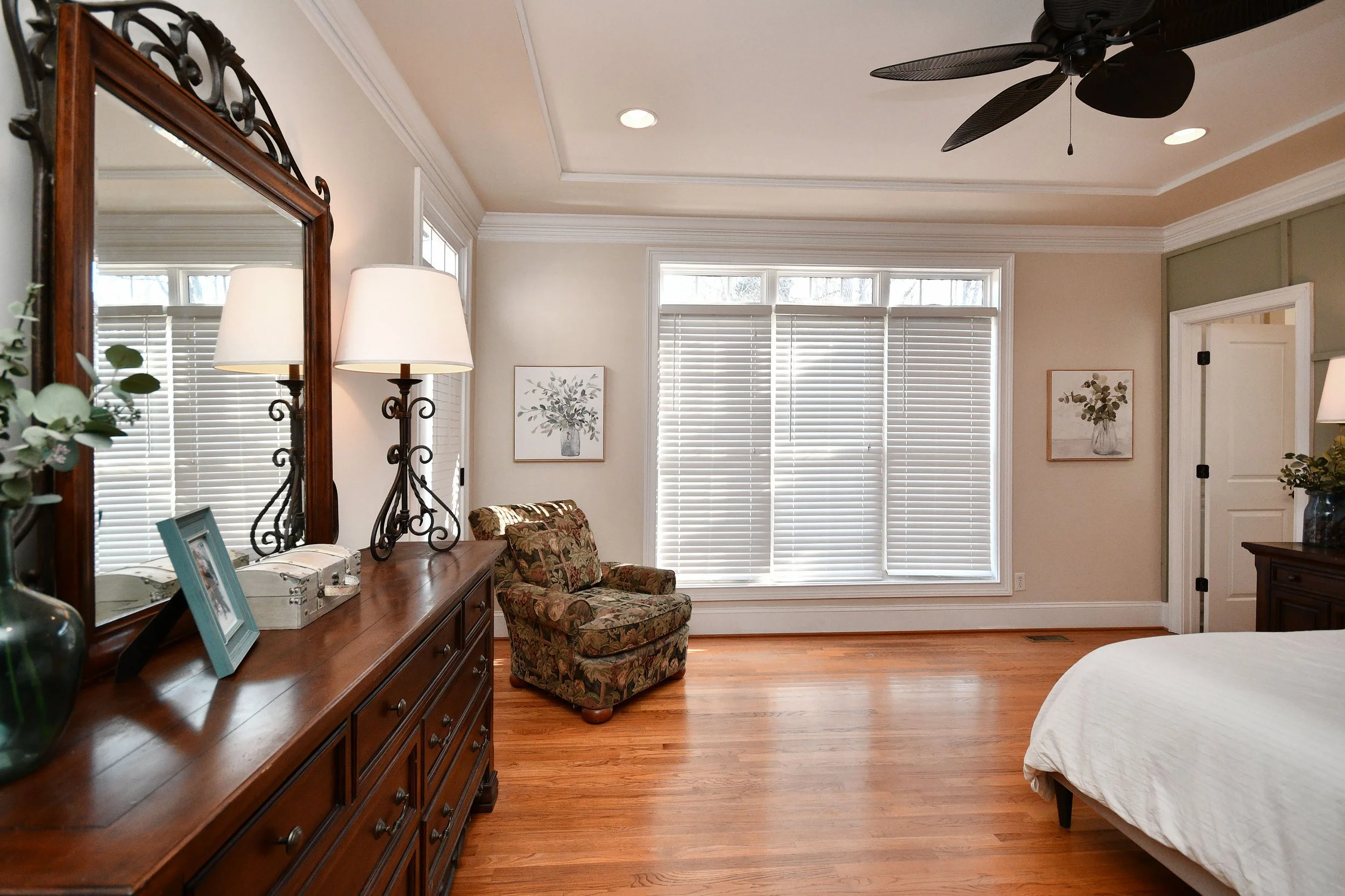 A bedroom with a wooden dresser, a mirror, lamps, a floral-patterned armchair, and framed artwork on the walls, with large windows and wooden floors. Home for sale in Greenville, SC. Master on Main Level. 4 Bedroom home for sale. Upscale Community. 