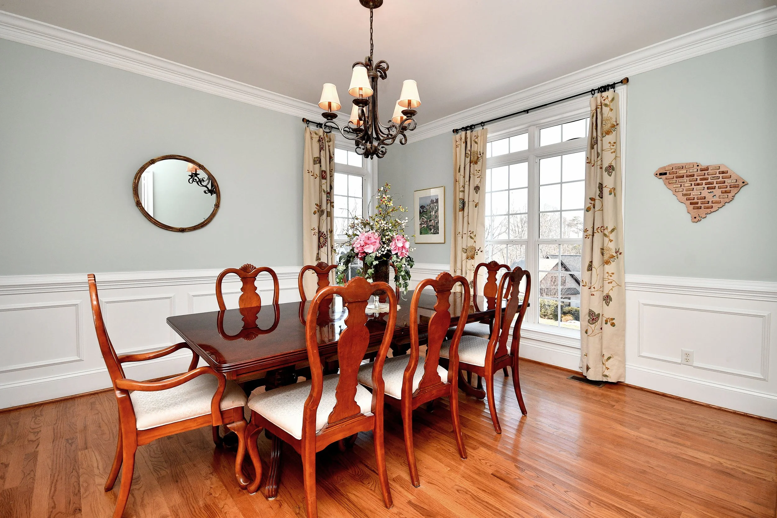 A dining room with a large table and eight matching chairs, a floral centerpiece, a chandelier hanging from the ceiling, large windows with floral curtains, a wall mirror, and decorative wall art. Home for sale in Greenville, SC. Master on Main.