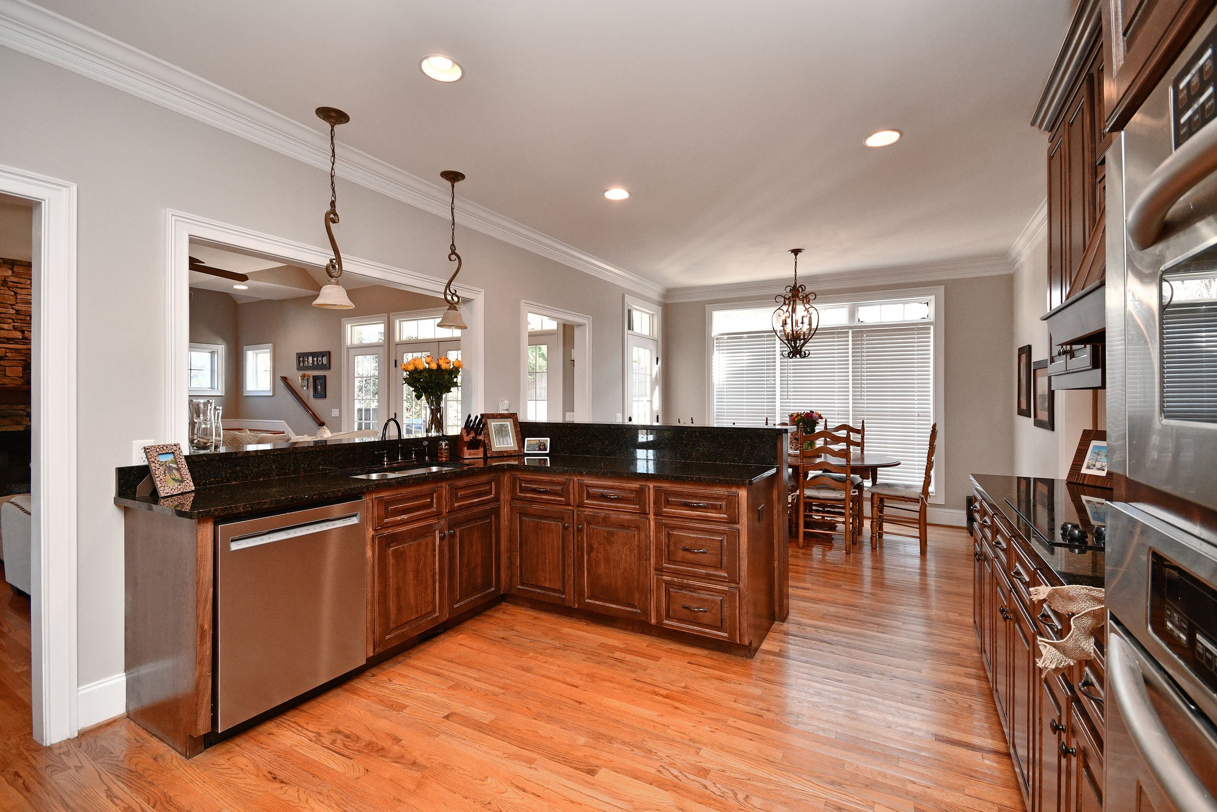 Kitchen with wooden cabinets, black countertops, stainless steel dishwasher, and a dining area with a round table and chairs near large windows with blinds. Home for sale in Greenville, SC. Master on Main Level. 4 Bedroom home for sale. Upscale 