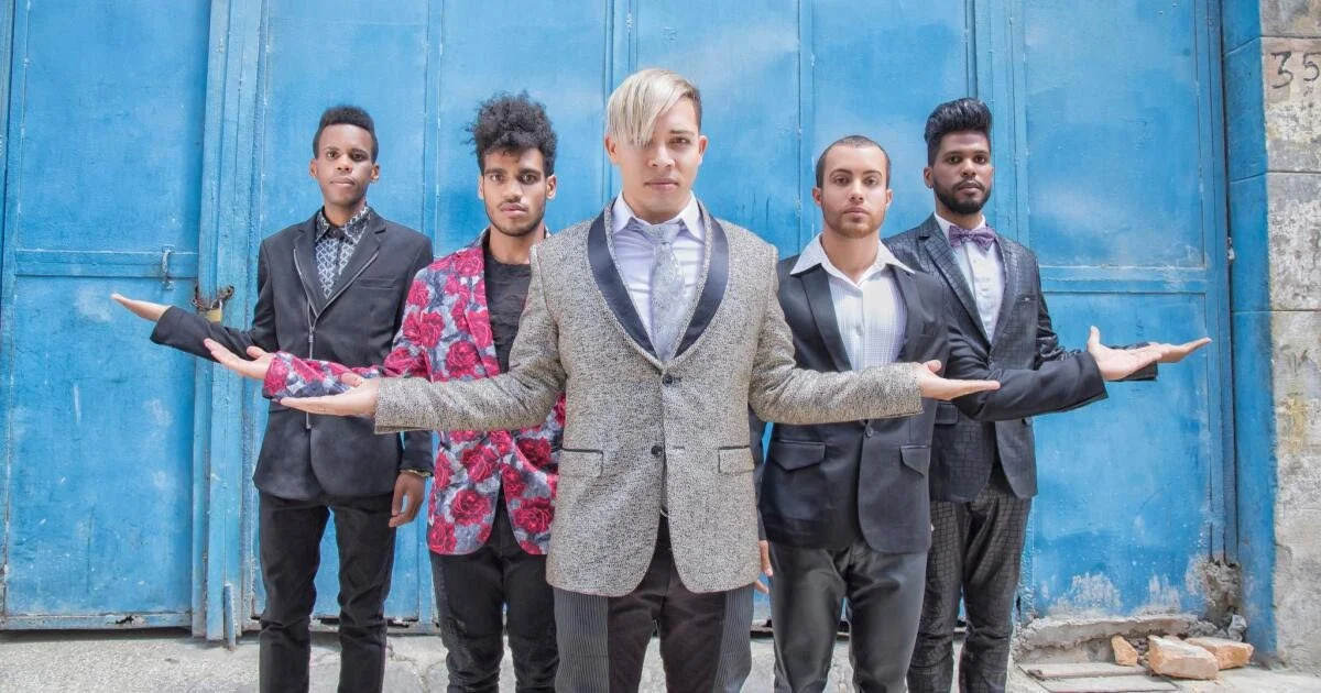 Five young men dressed in fashionable suits and jackets standing in front of a textured blue metal door with their arms extended sideways.