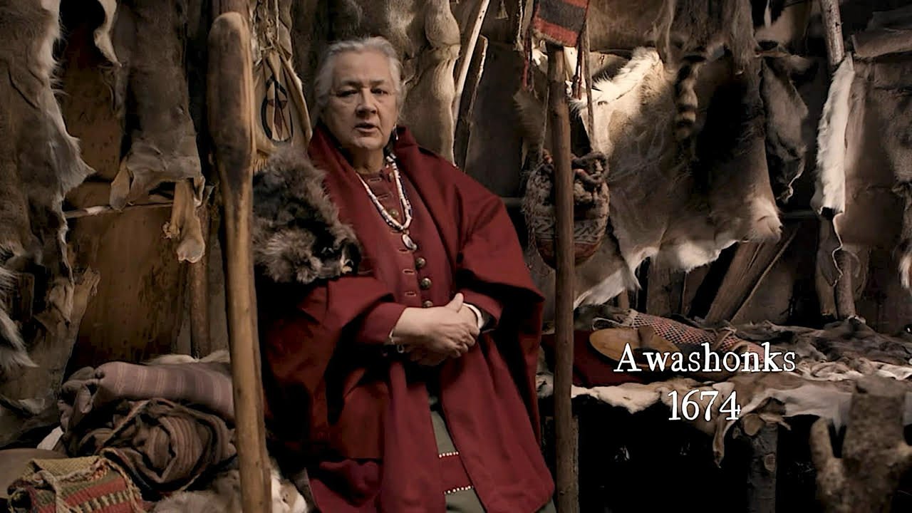 A person dressed in traditional Native American clothing standing in front of fur hides and animal pelts with the text 'Awashonks 1674' displayed.
