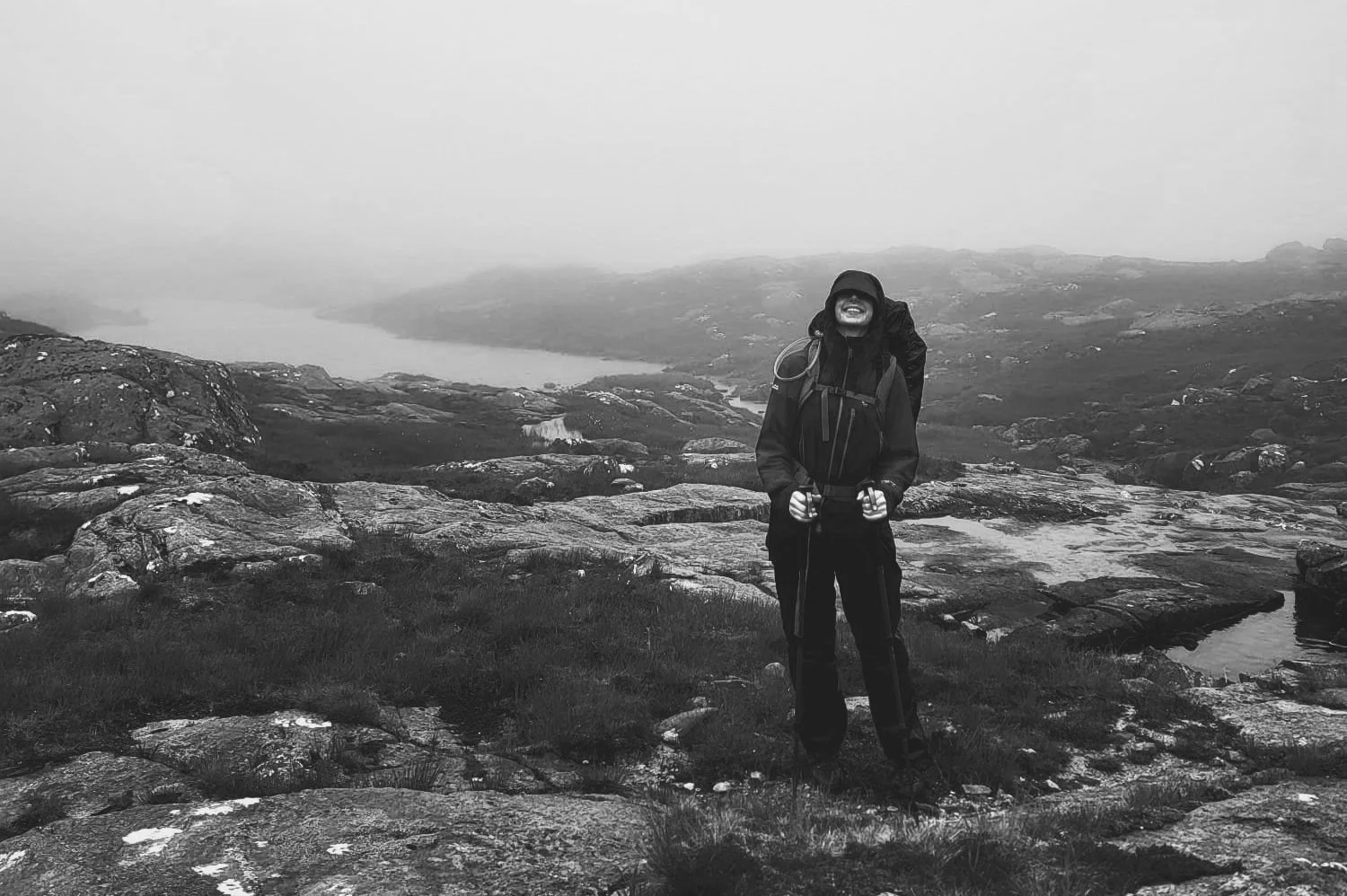 A person dressed in outdoor hiking gear with a backpack, standing on rocky terrain in a mountainous area under a cloudy sky, smiling, with a distant body of water and hills in the background.