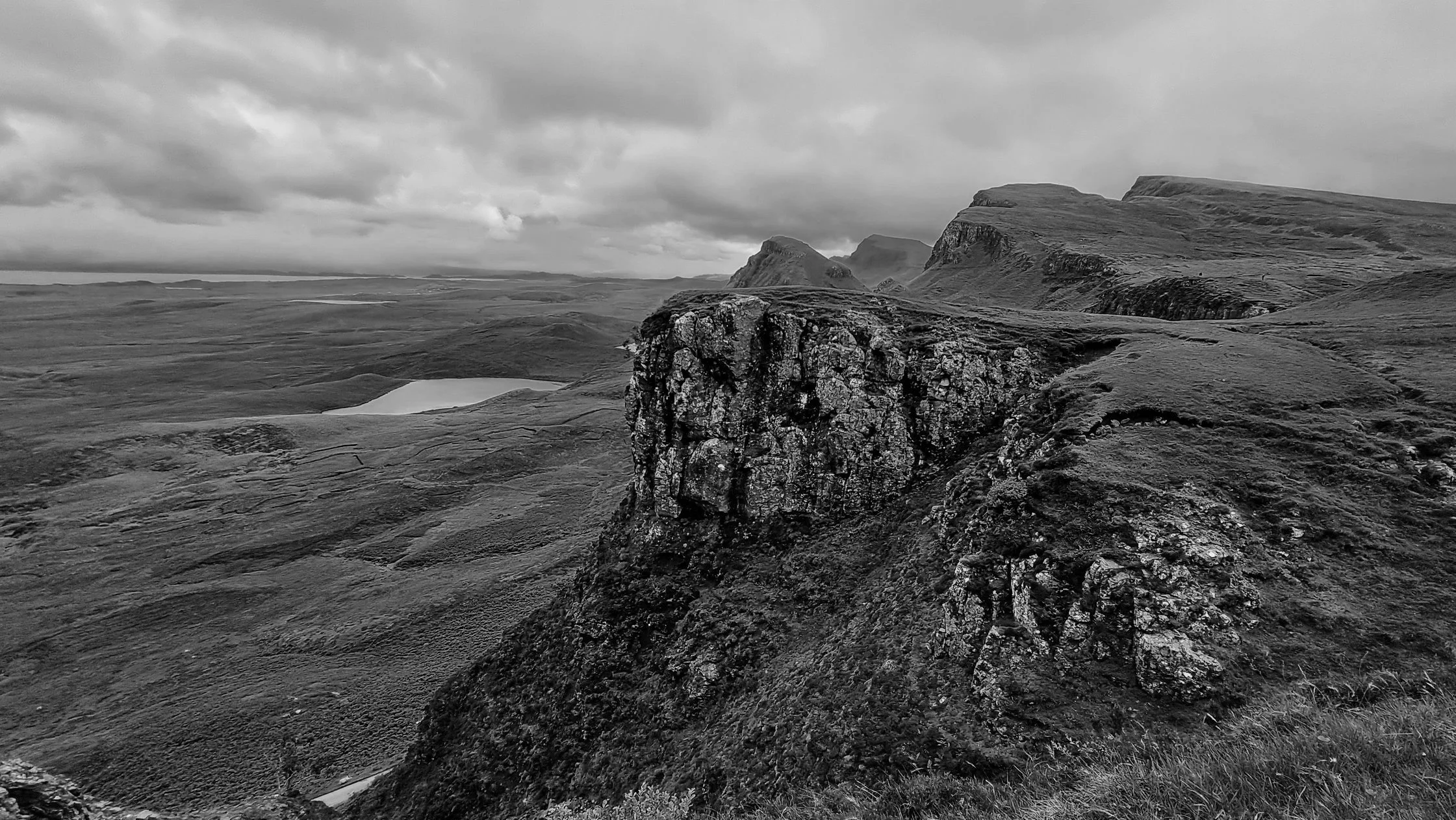 Scenic black and white landscape with rolling hills, a rocky prominence in the foreground, and distant mountains under a cloudy sky.