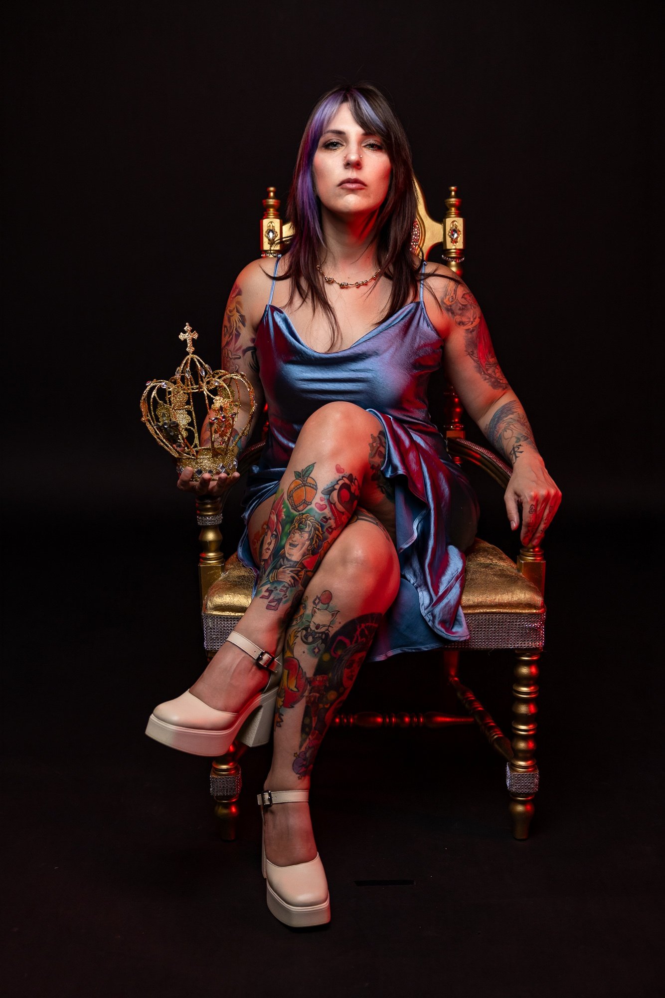 Woman with tattoos sitting on a throne holding a crown, wearing a blue dress and white heels, against a black background.