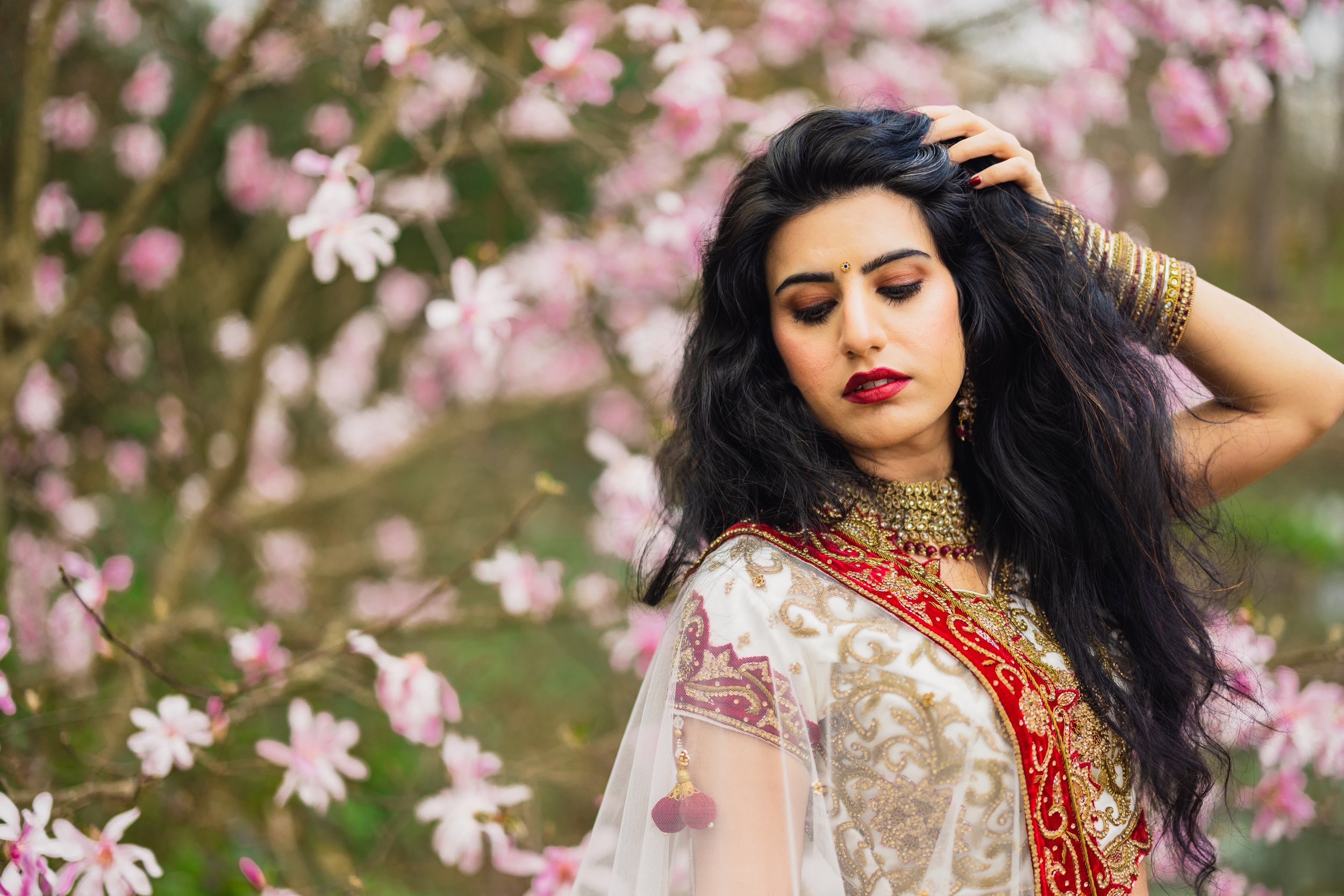 Woman in traditional Indian attire posing near blooming cherry blossoms, wearing a white and red embellished sari with gold jewelry and long dark hair.