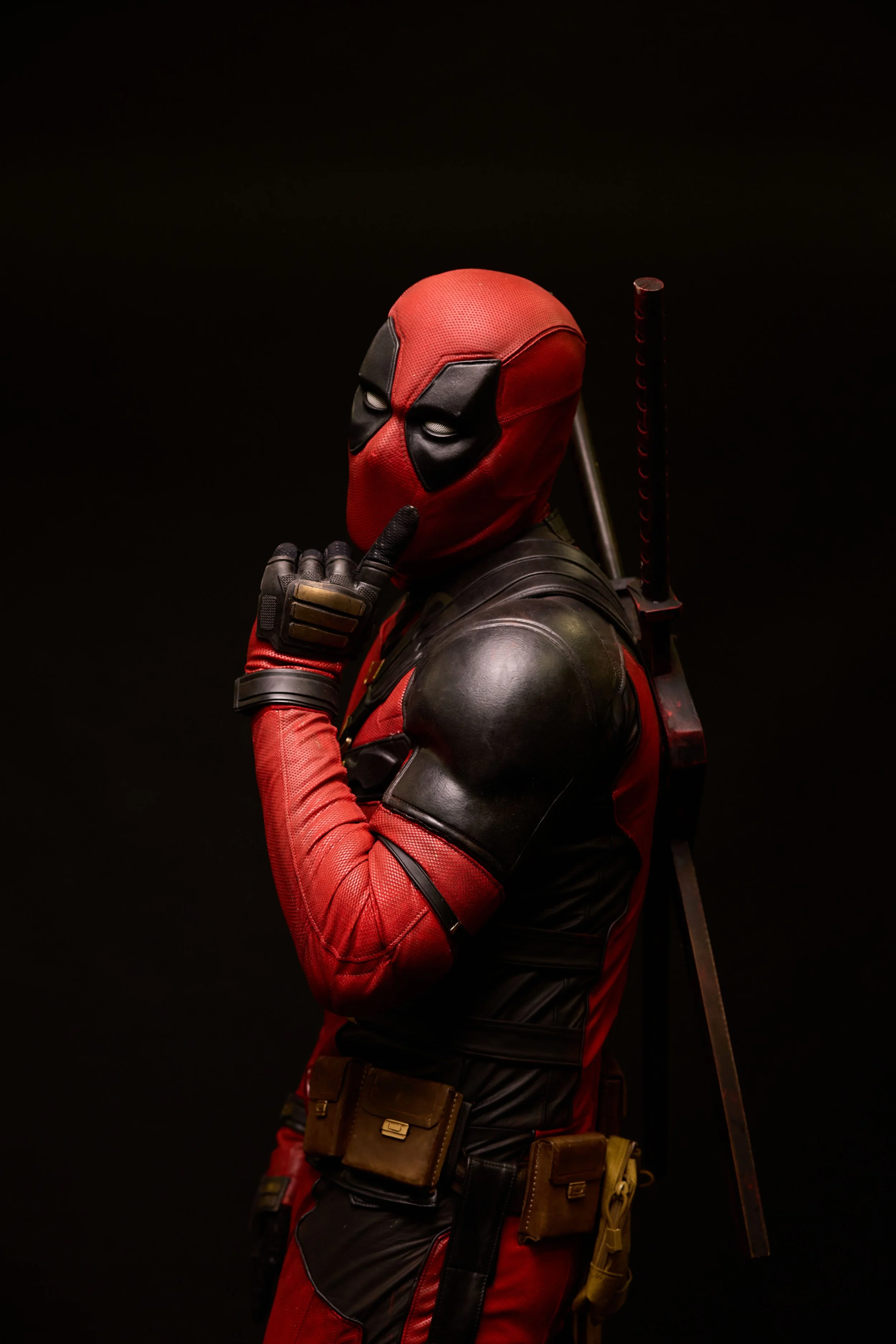Superhero in red and black costume with finger on lips, holding a sword on the back, against a dark background.