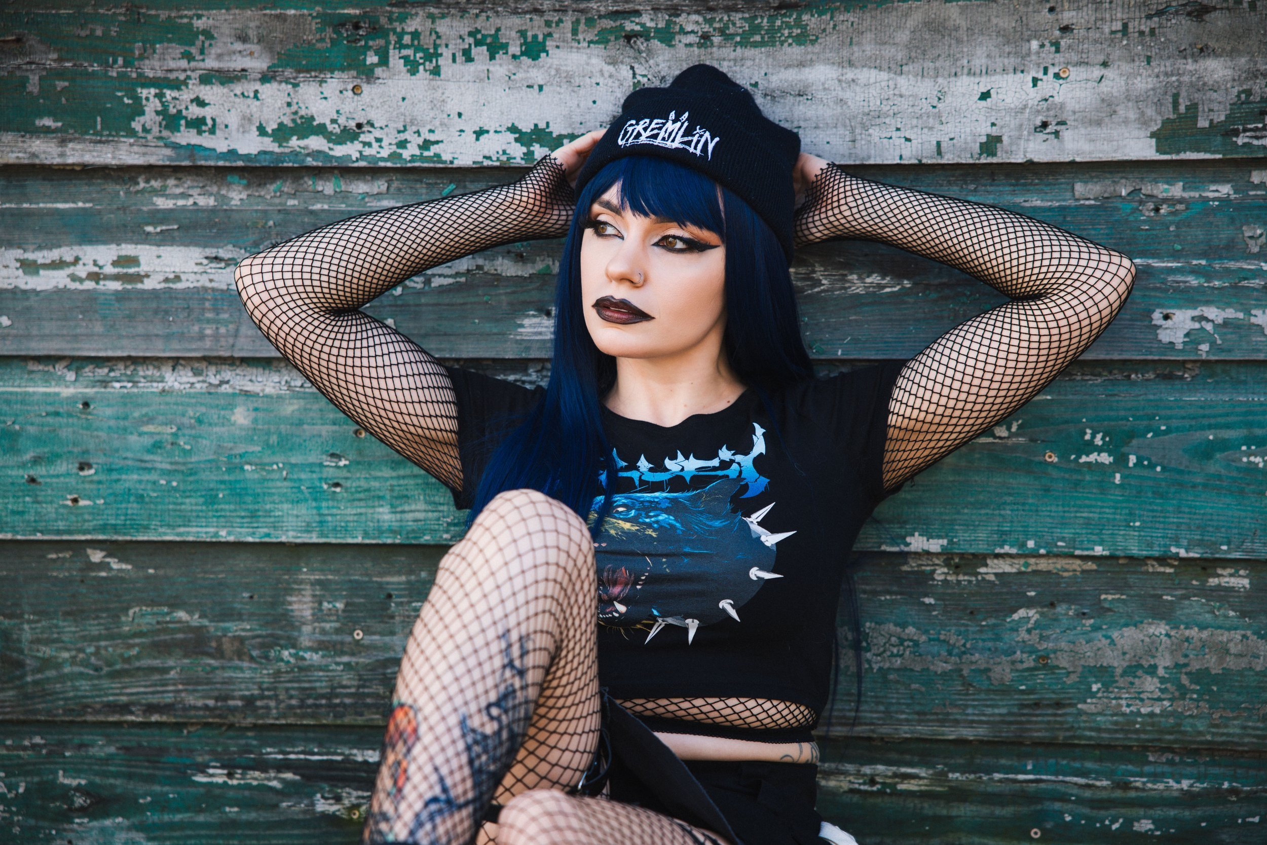 Person in a black beanie and fishnet outfit sitting against a weathered wooden wall