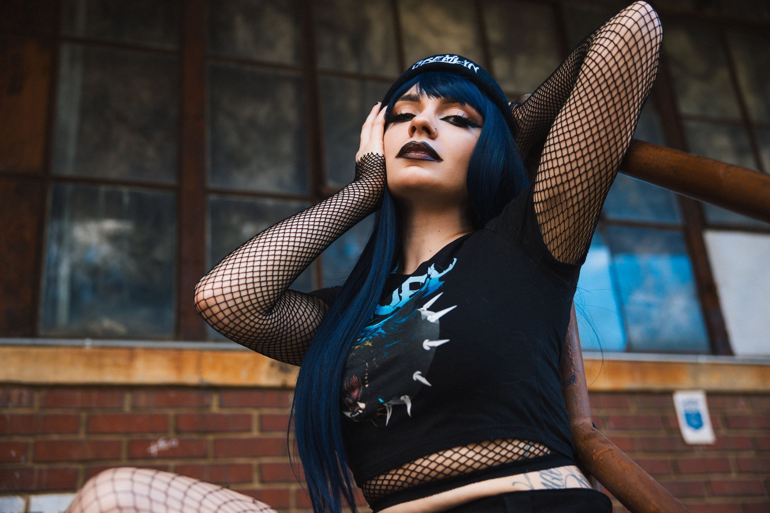 Woman with blue hair wearing fishnet sleeves and dark lipstick, sitting on stairs in front of a brick building.