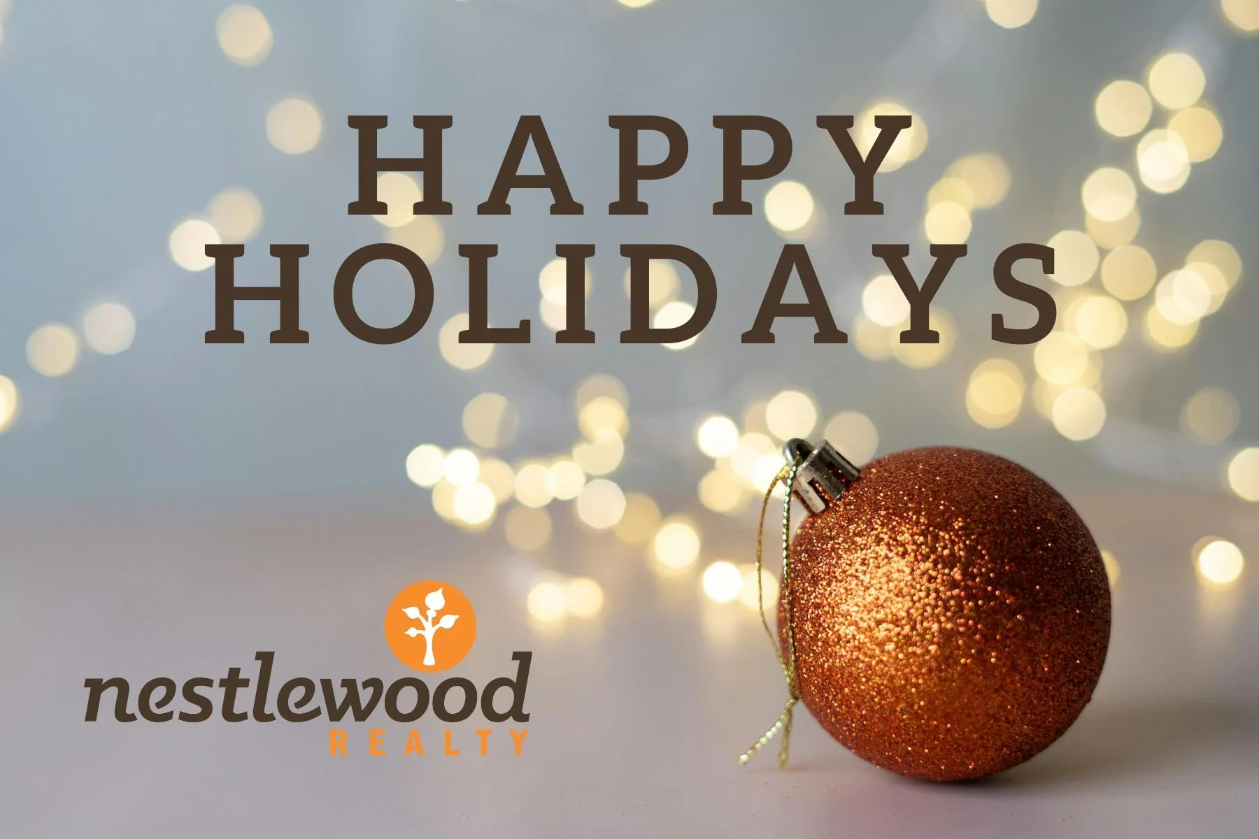 A festive holiday greeting card with the text 'HAPPY HOLIDAYS' at the top, a shiny orange Christmas ornament at the bottom right, and the Nestlewood Realty logo at the bottom left, with a background of blurred warm white string lights.
