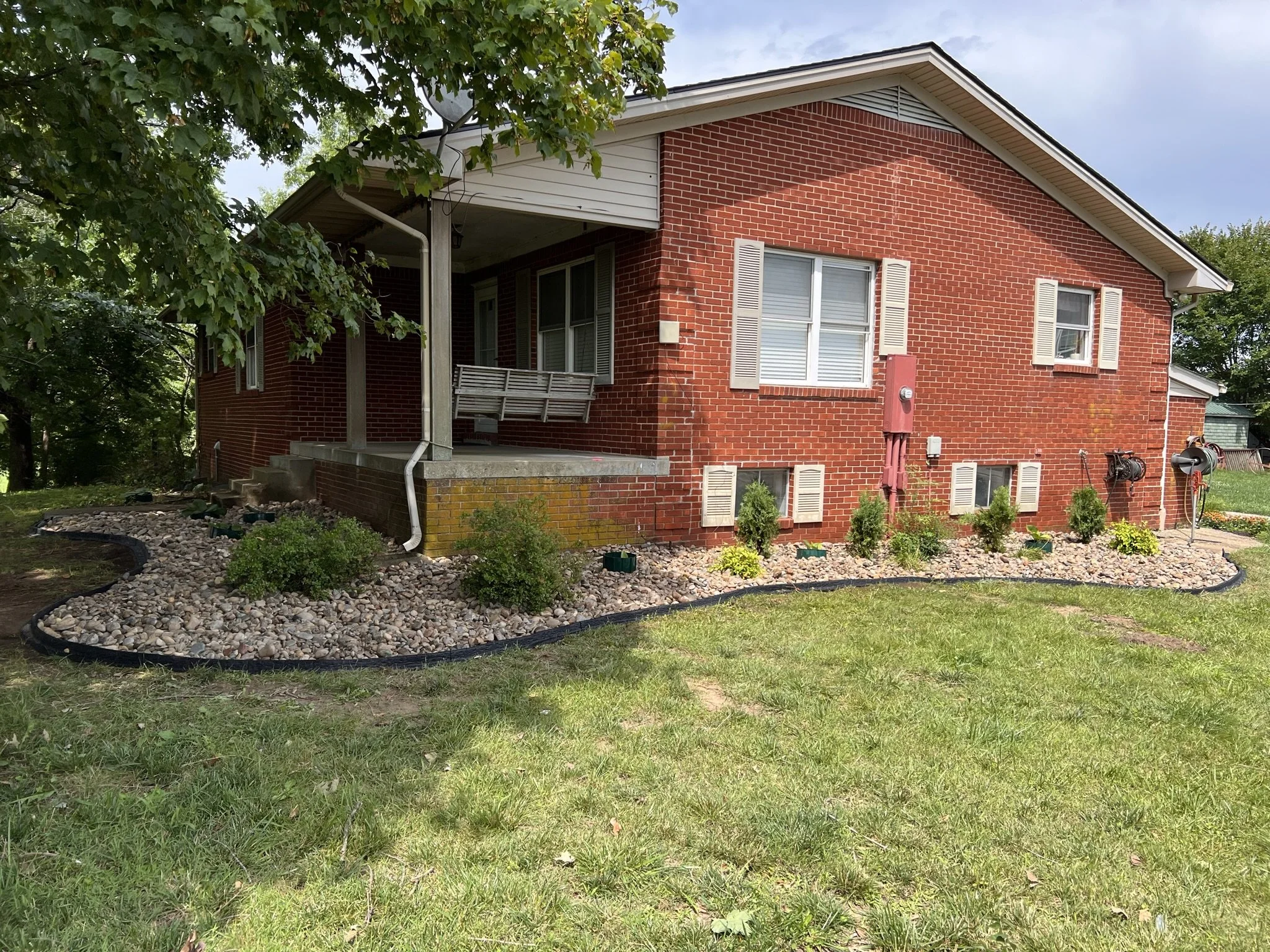A red brick house with white shutters and a front porch with a swing, surrounded by a landscape of small bushes and rocks, with a tree on the left and a grassy yard.