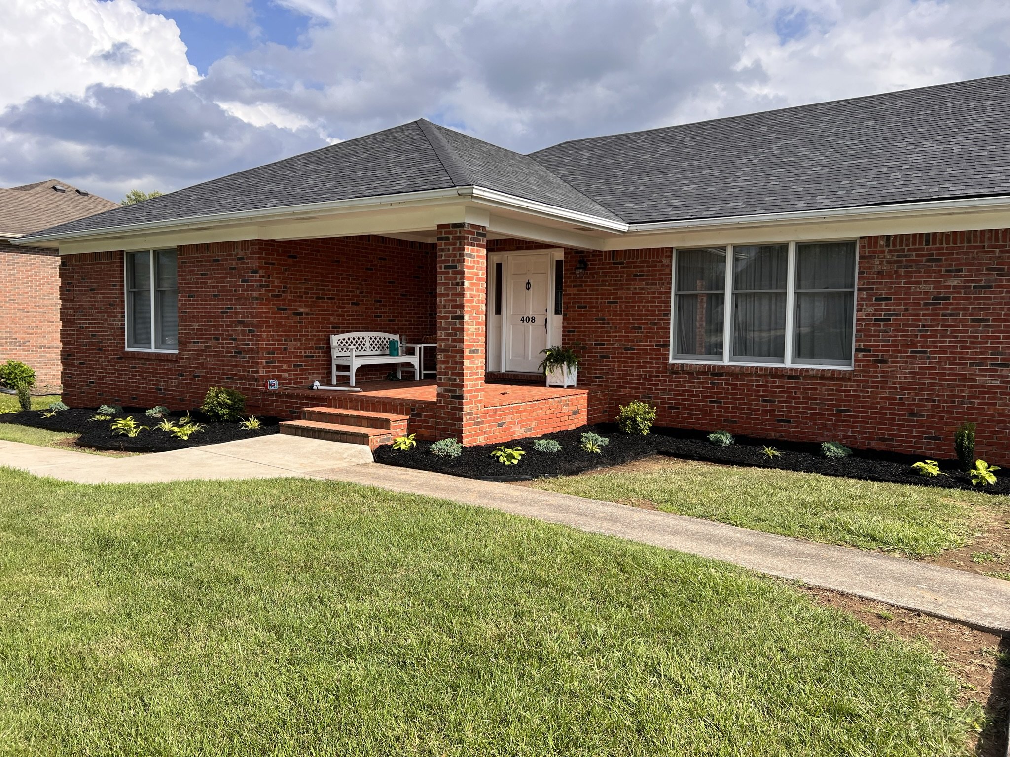 A brick house with a porch, white bench, front steps, and a lawn with freshly planted garden beds.