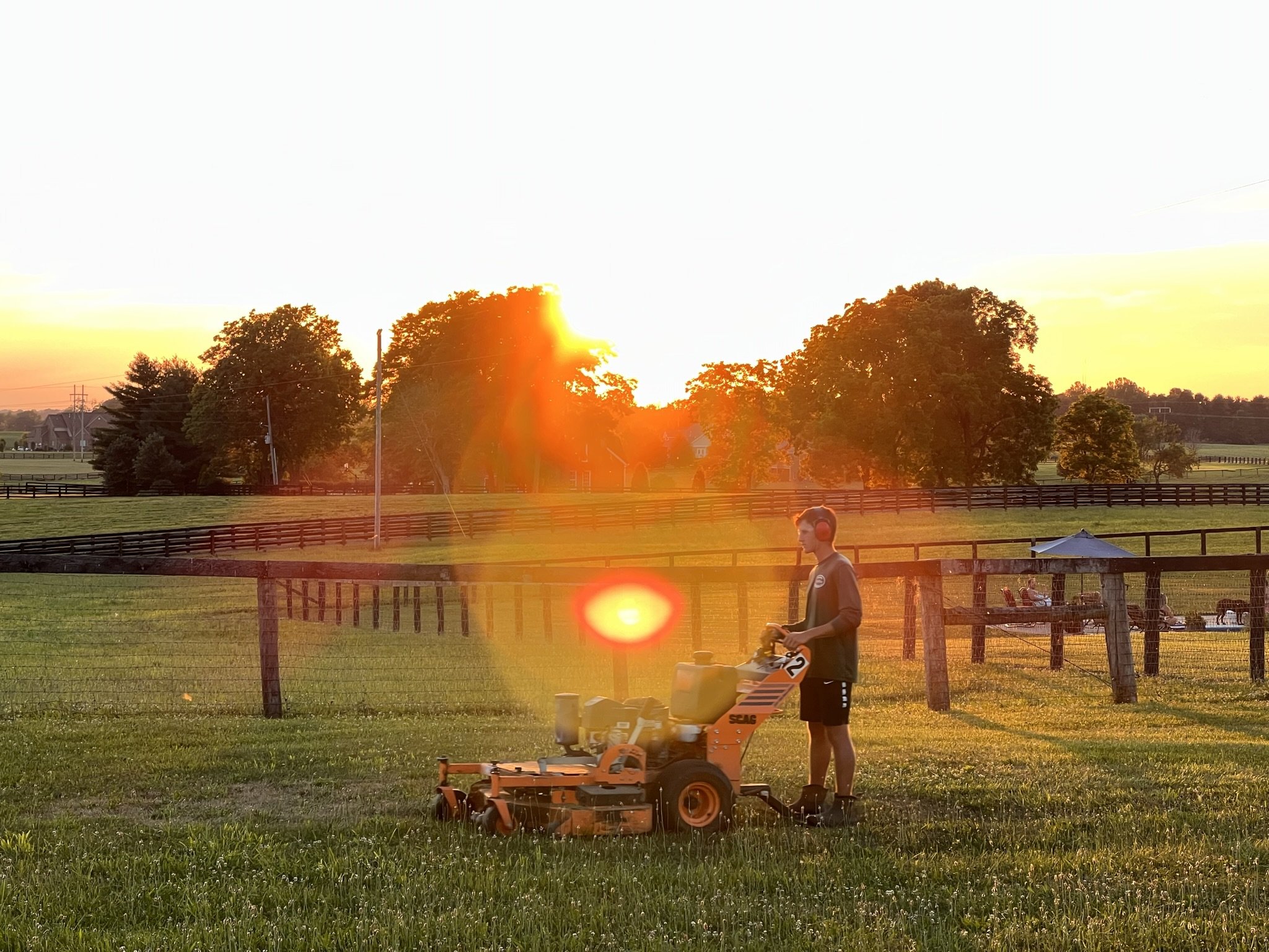 A person wearing headphones operating a mower on a grassy field during sunset, with trees and a fenced pasture in the background.