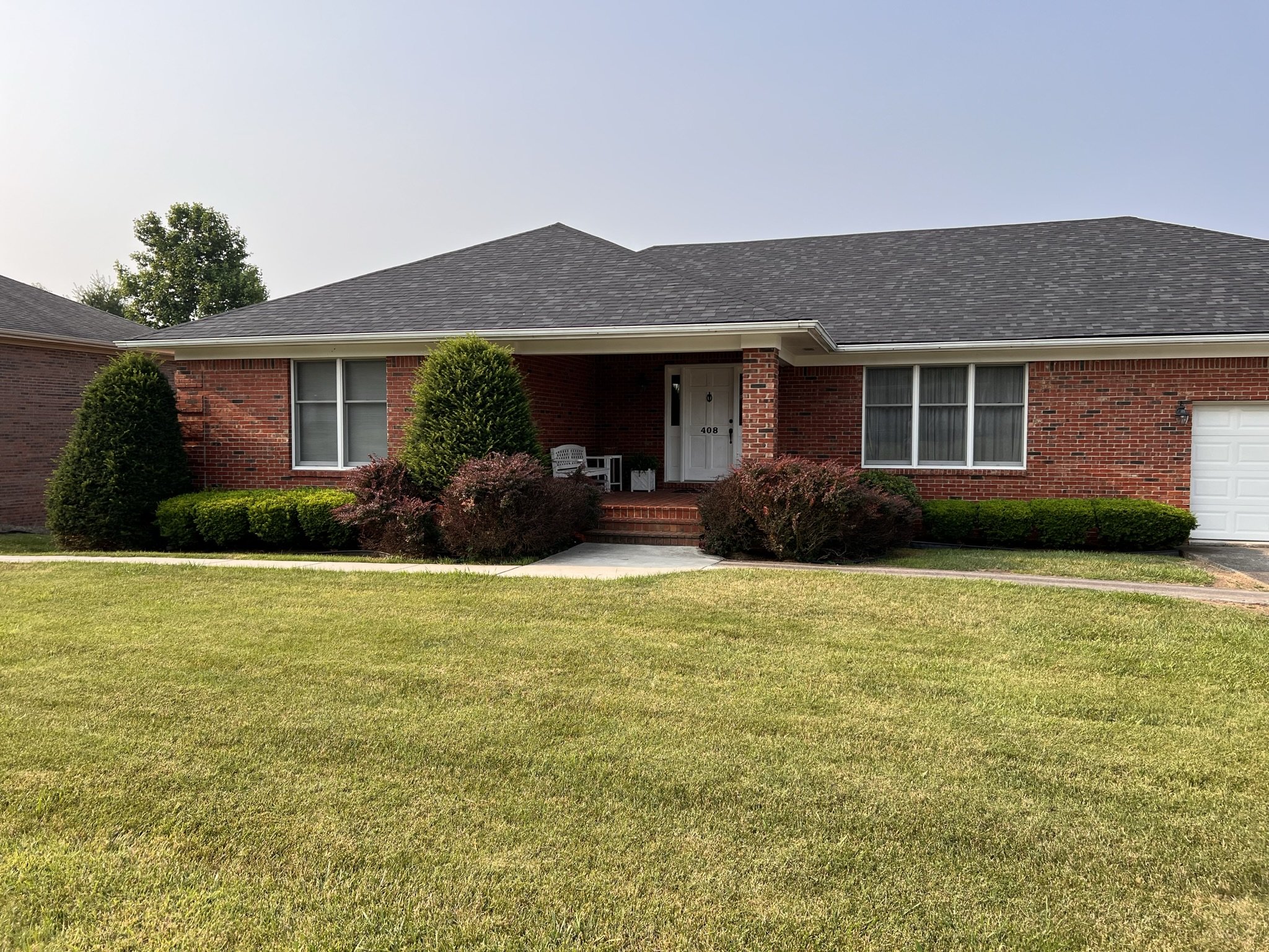 A single-story red brick house with a gray shingled roof, white garage door, front porch, and well-maintained front yard with green grass and bushes.