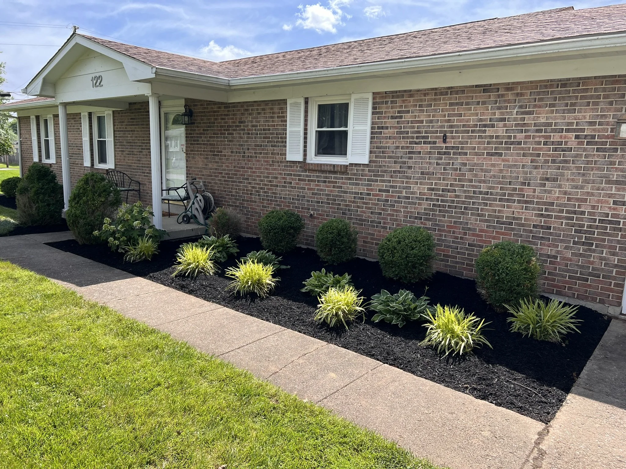 Front yard garden with small bushes, plants, mulch, and a sidewalk in front of a brick house with a porch, windows, and a grass lawn.