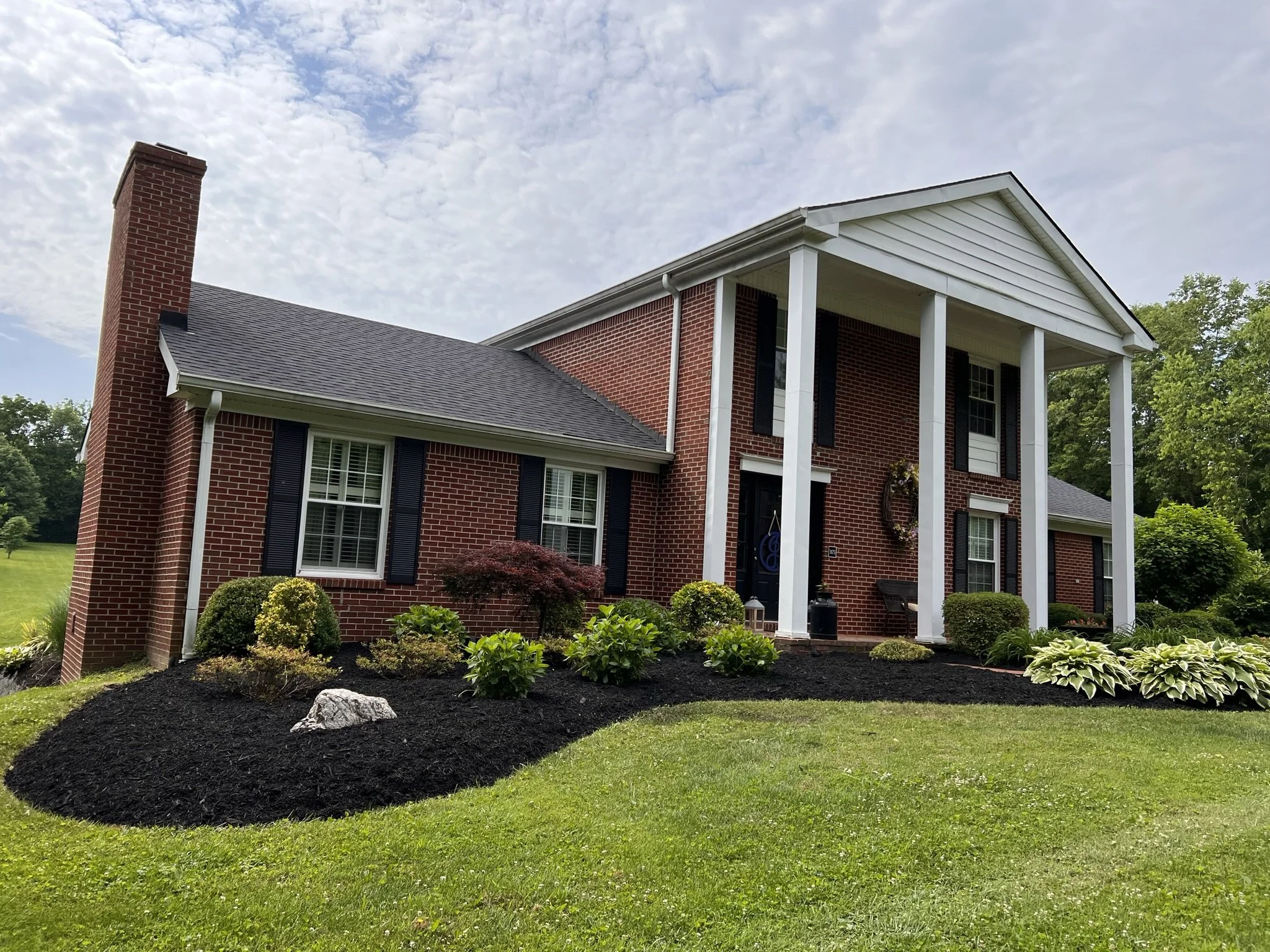Front view of a two-story brick house with white columns and black shutters, surrounded by a well-manicured lawn and landscaped garden with various bushes and plants.