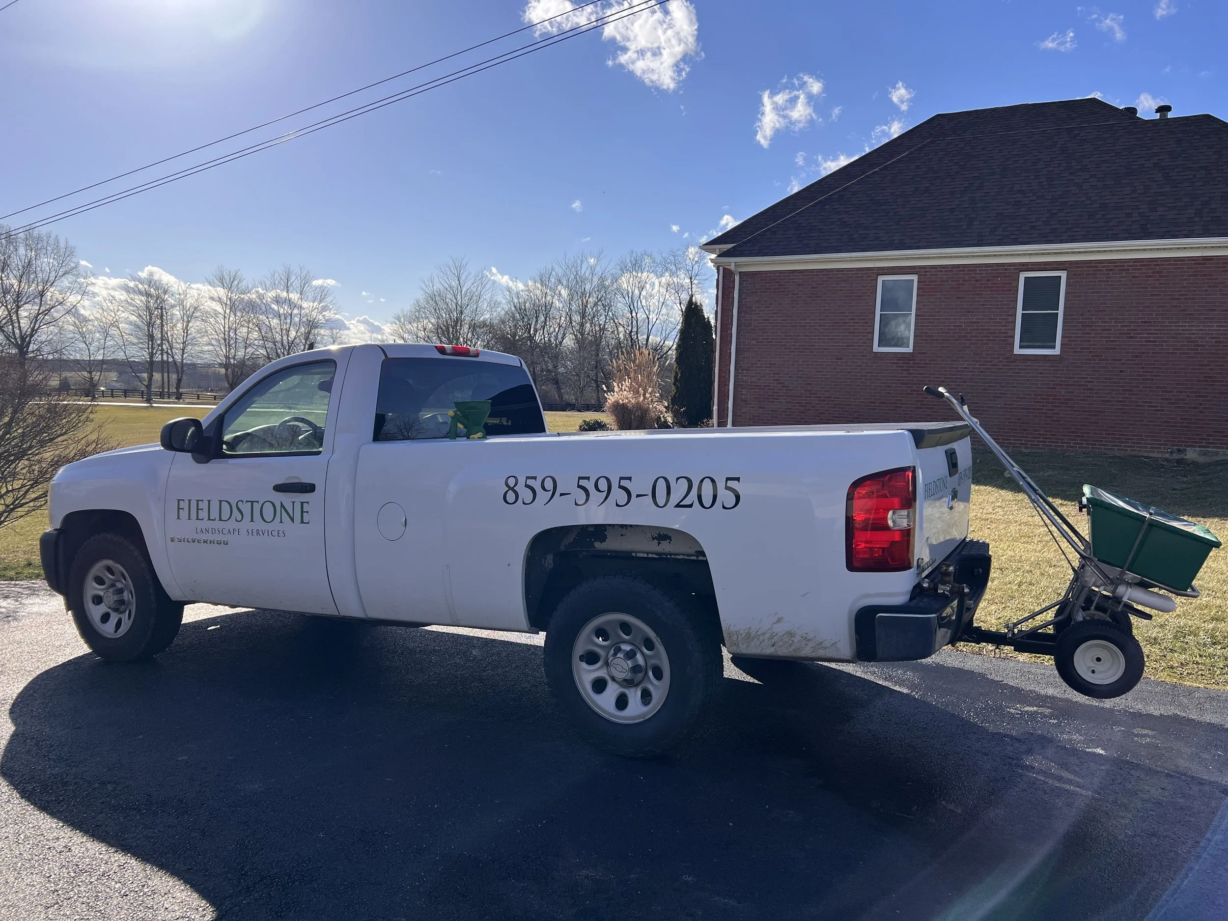 White pickup truck with landscaping company branding, parked on a driveway near a brick house, with a green garden cart attached to the back.