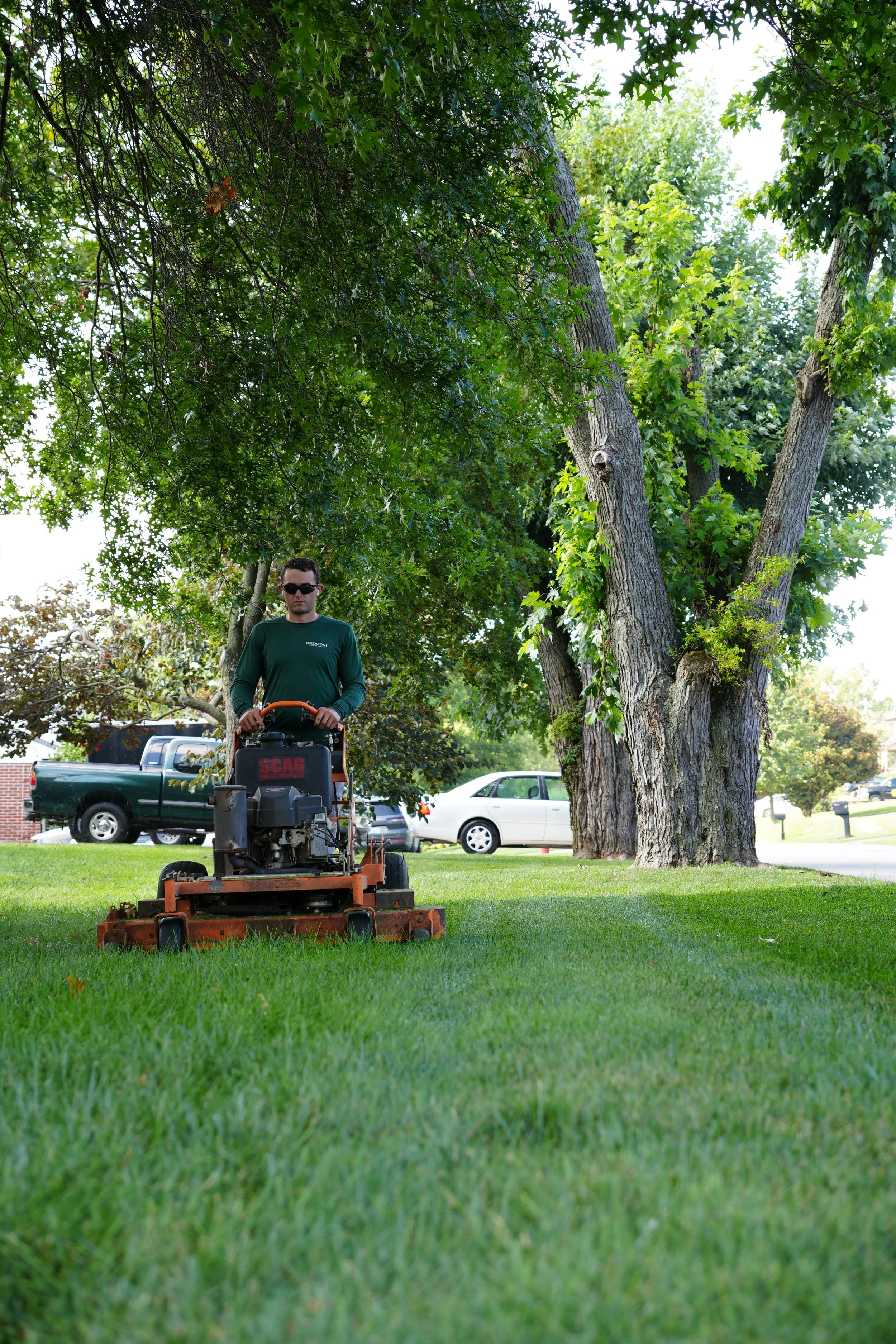 A man wearing sunglasses and a dark green long-sleeve shirt mowing a lawn with a riding mower under a large leafy tree in a residential neighborhood.