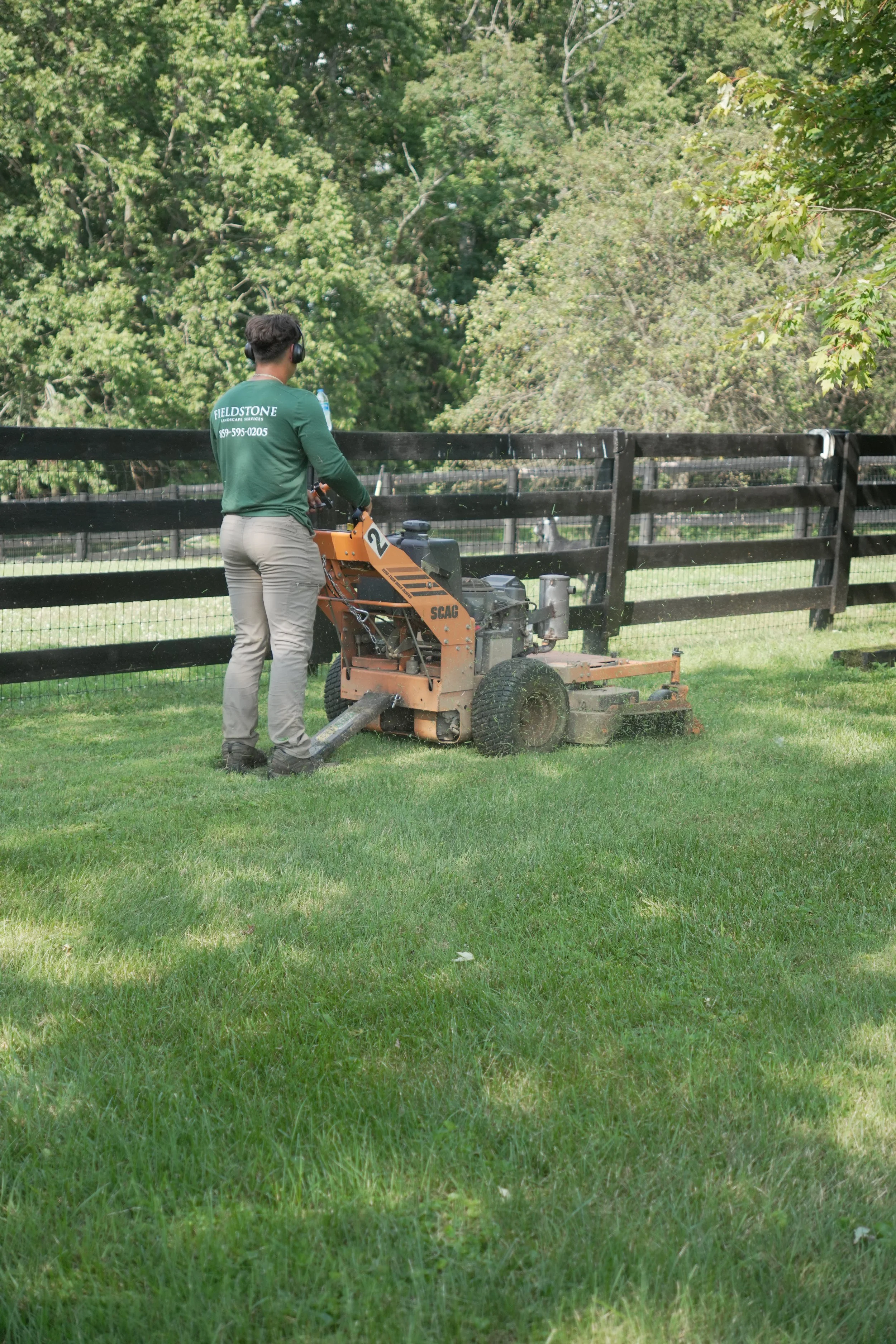 A person operating a line-trimmer in a green backyard, maintaining the grass near a black wooden fence surrounded by trees.