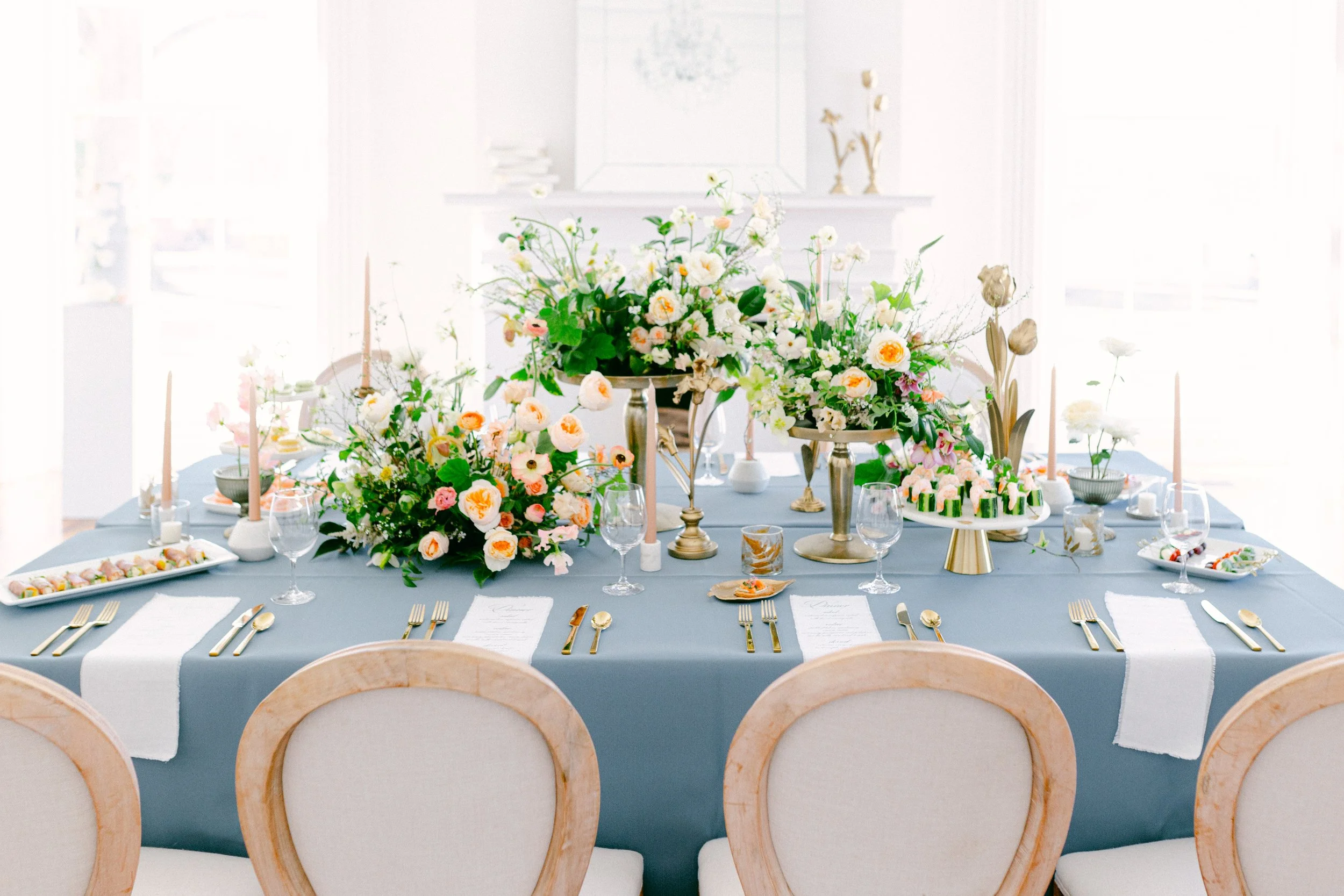 Elegant table setting with floral arrangements, blue tablecloth, decorative plates, gold cutlery, and candles. Photo used to give viewer event styling inspiration.
