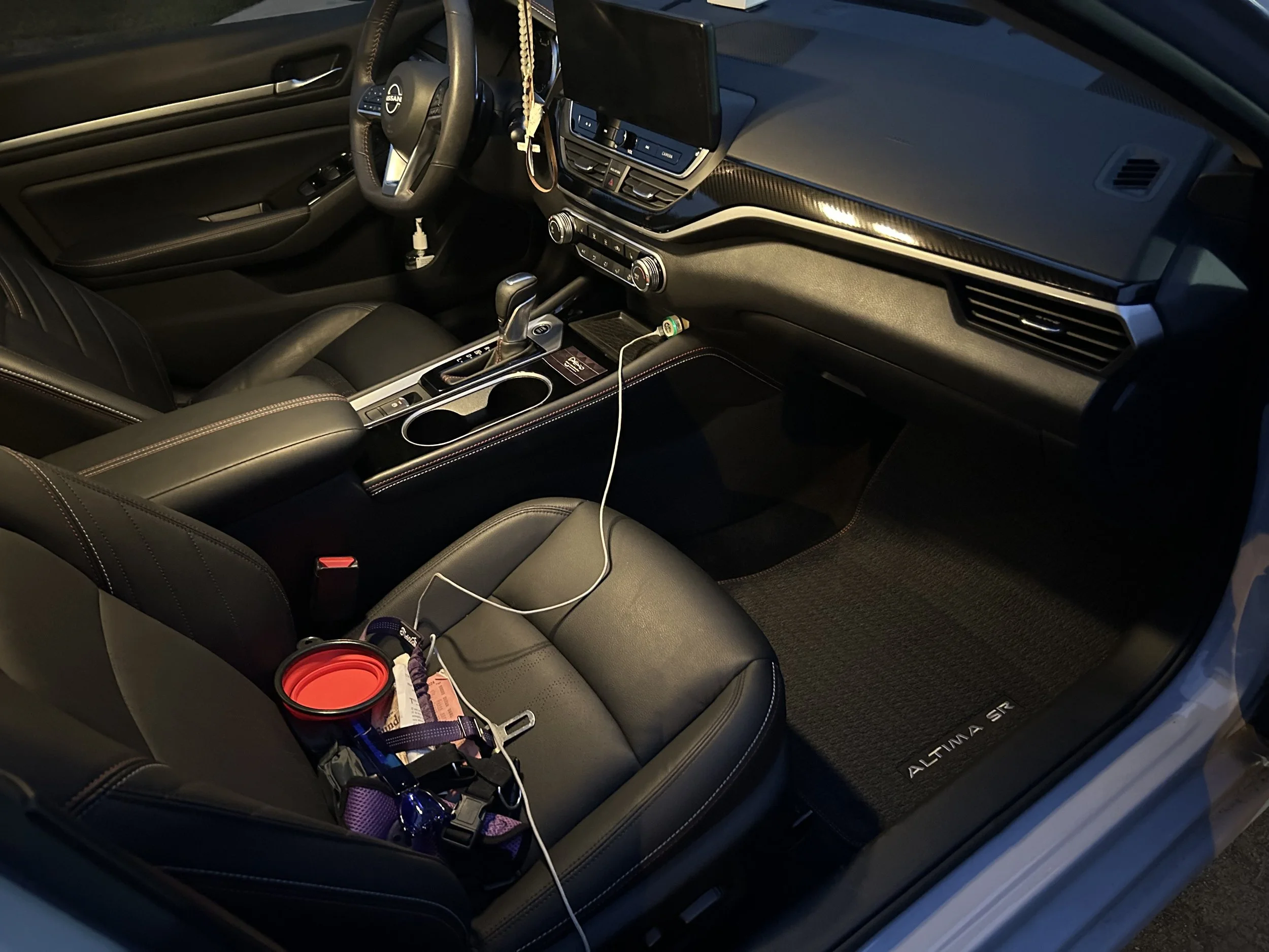 Interior of a Nissan Altima SR with leather seats, console, and steering wheel, decorated with personal items like a phone charger and pet accessories on the seat.