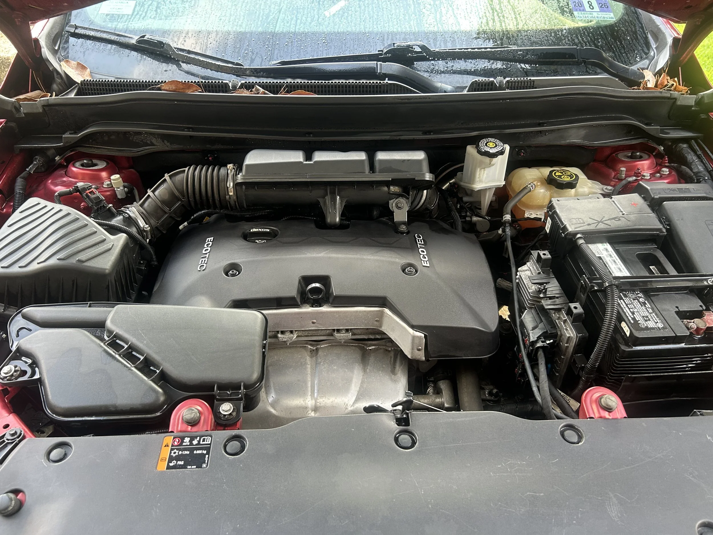 A clean car engine compartment with a black EcoTec engine cover, surrounding components, and a windshield with rain droplets visible.