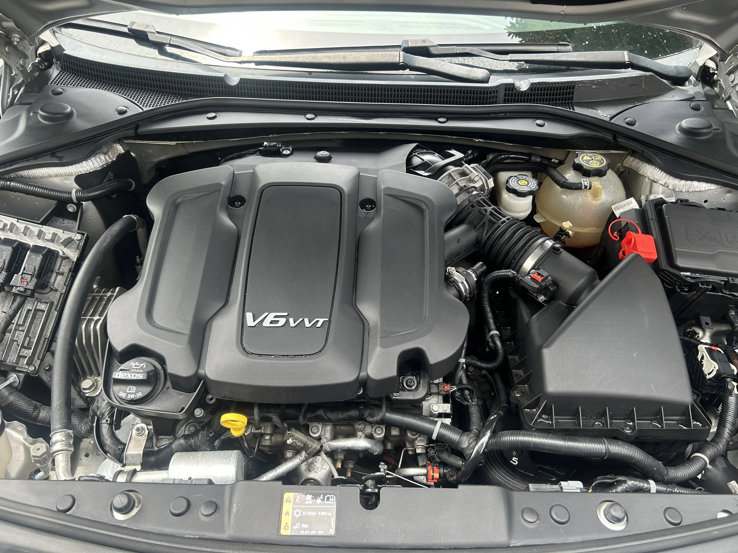 Front view of a car engine bay with a V6 VVT engine cover, various hoses, fluid reservoirs, and a battery.