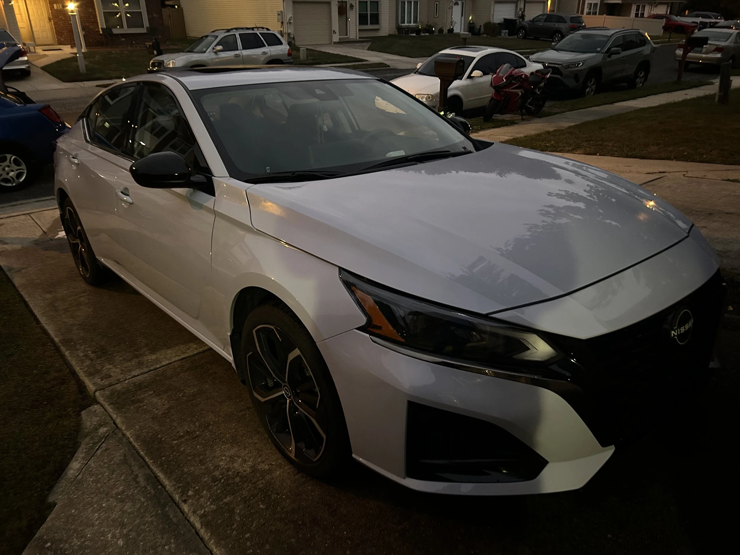 White Nissan sedan parked in a residential driveway at dusk, surrounded by other cars and motorcycles.