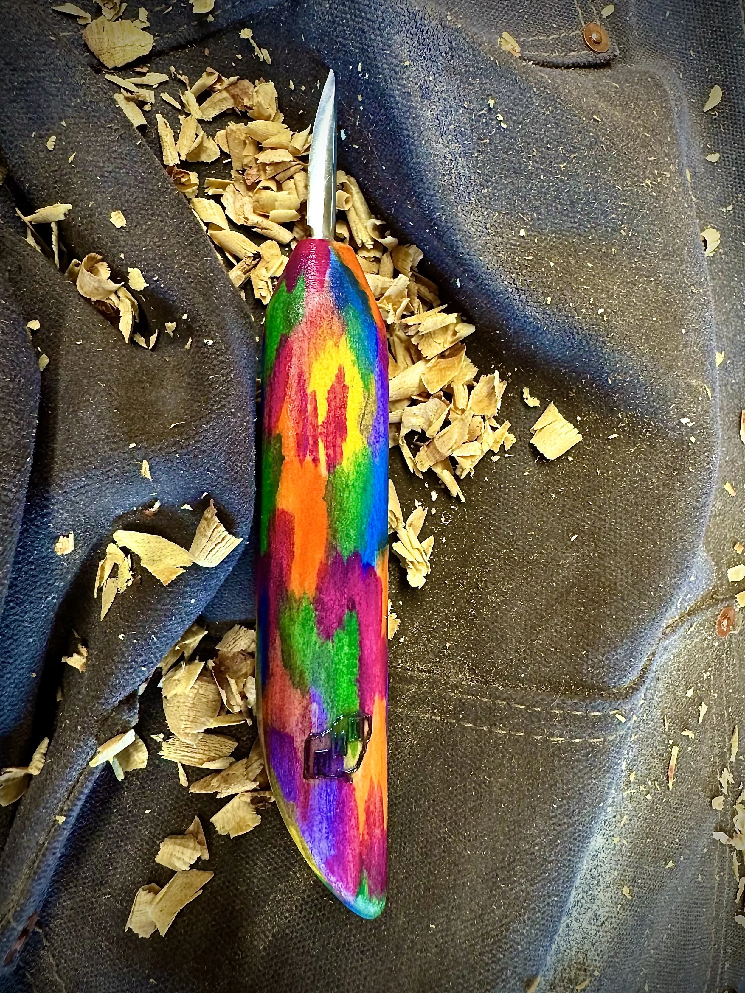 A colorful woodworking chisel with a rainbow painted handle, placed on a dark surface with wood shavings around it.