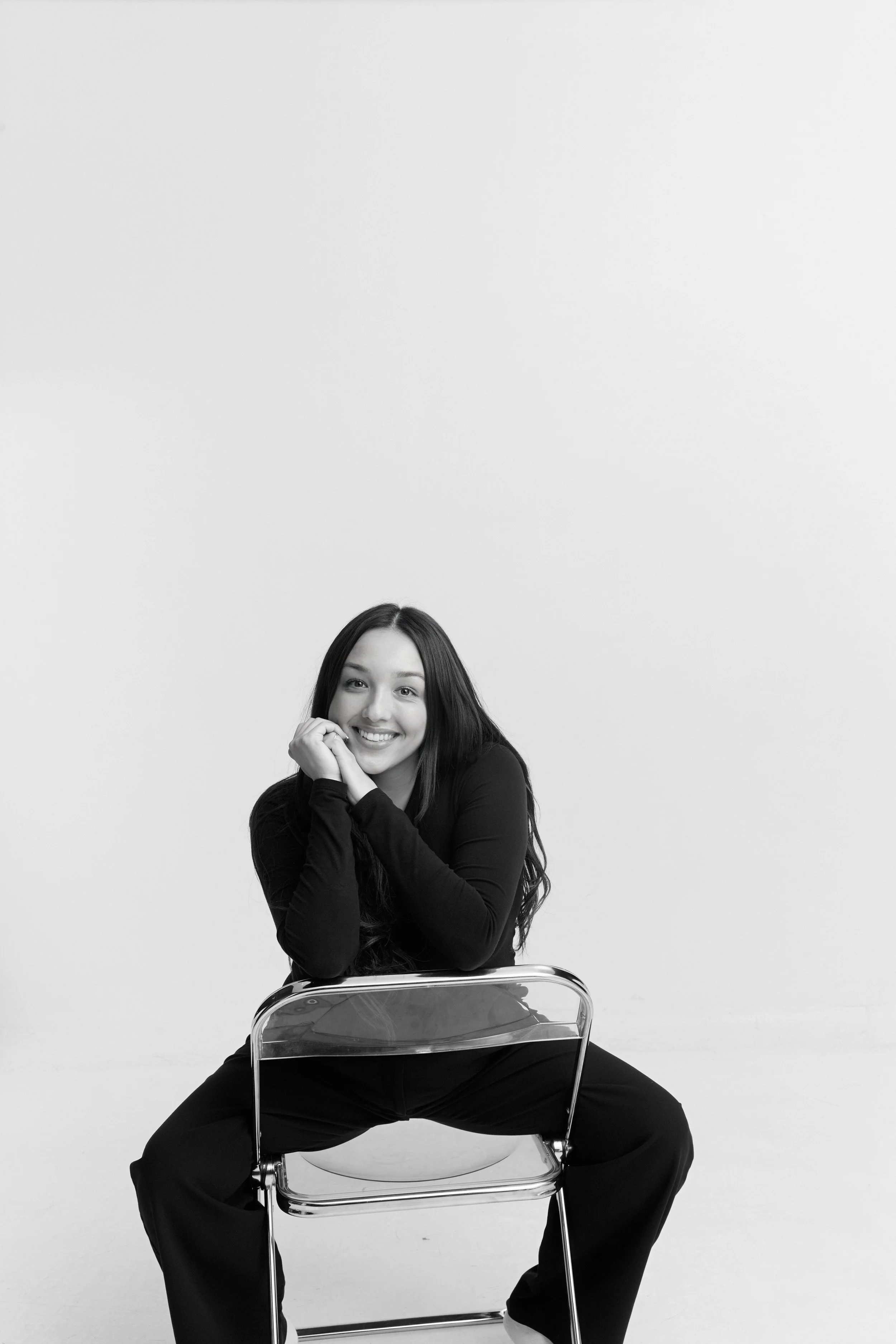 Black and white studio portrait of a smiling young woman with long hair, sitting on a transparent chair, wearing a long-sleeve top and Pants, with her hands near her face, and legs spread apart.