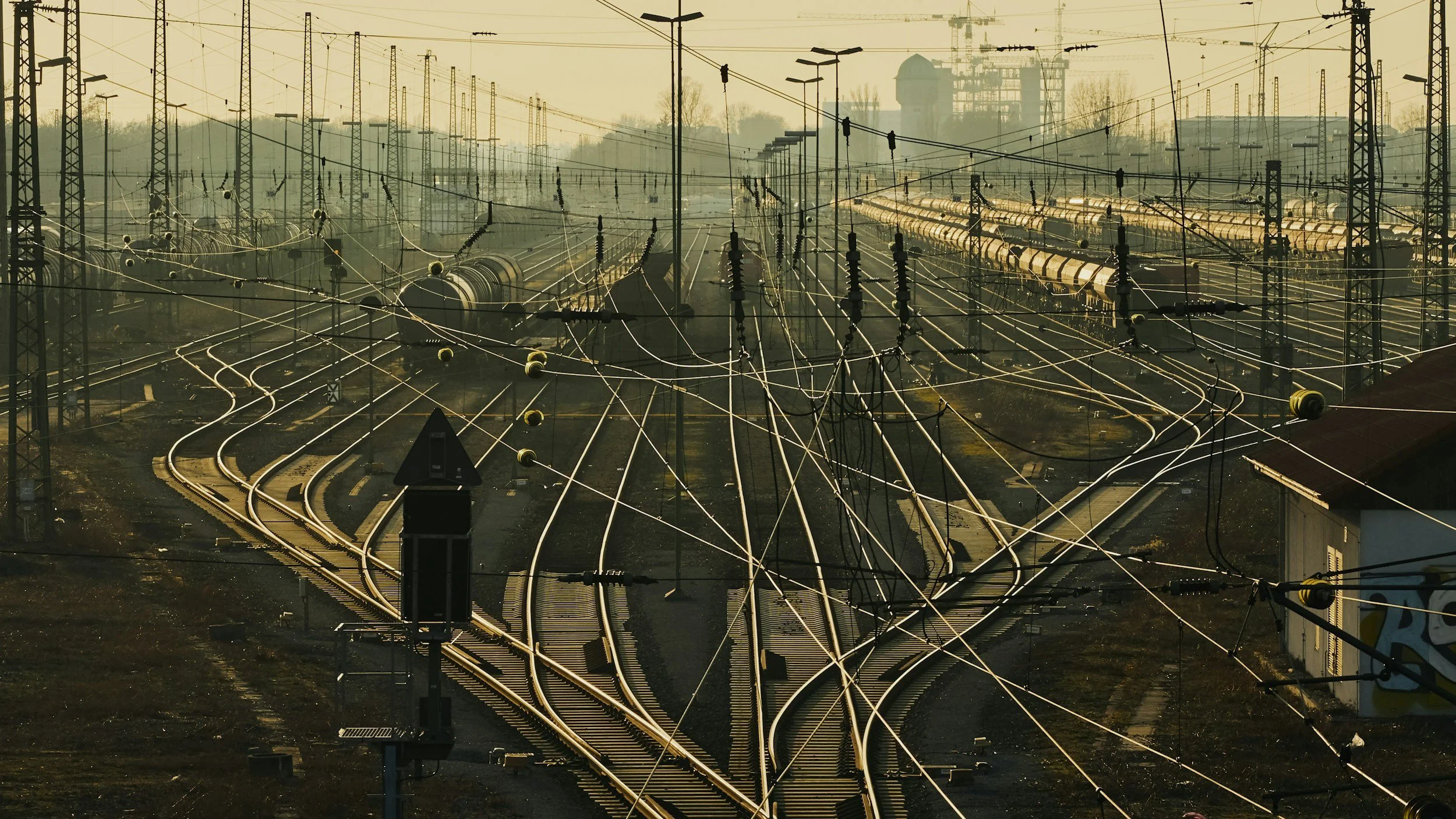 A network of train tracks with overhead electrical lines and pylons, under a hazy, muted sky.