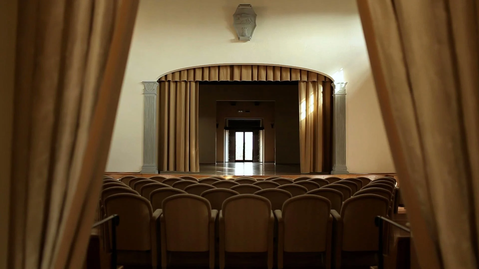 View of an empty theater or auditorium from behind golden curtains, showing rows of chairs facing a stage with a closed door in the background, framed by ornate columns and a curved valance above.