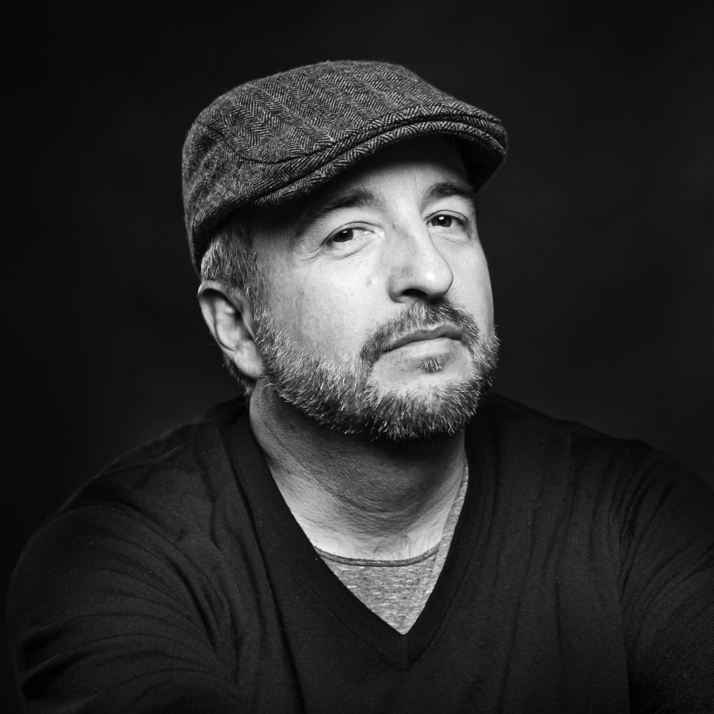 Black and white photo of a man with a beard wearing a flat cap, gray shirt, and black jacket, against a dark background.