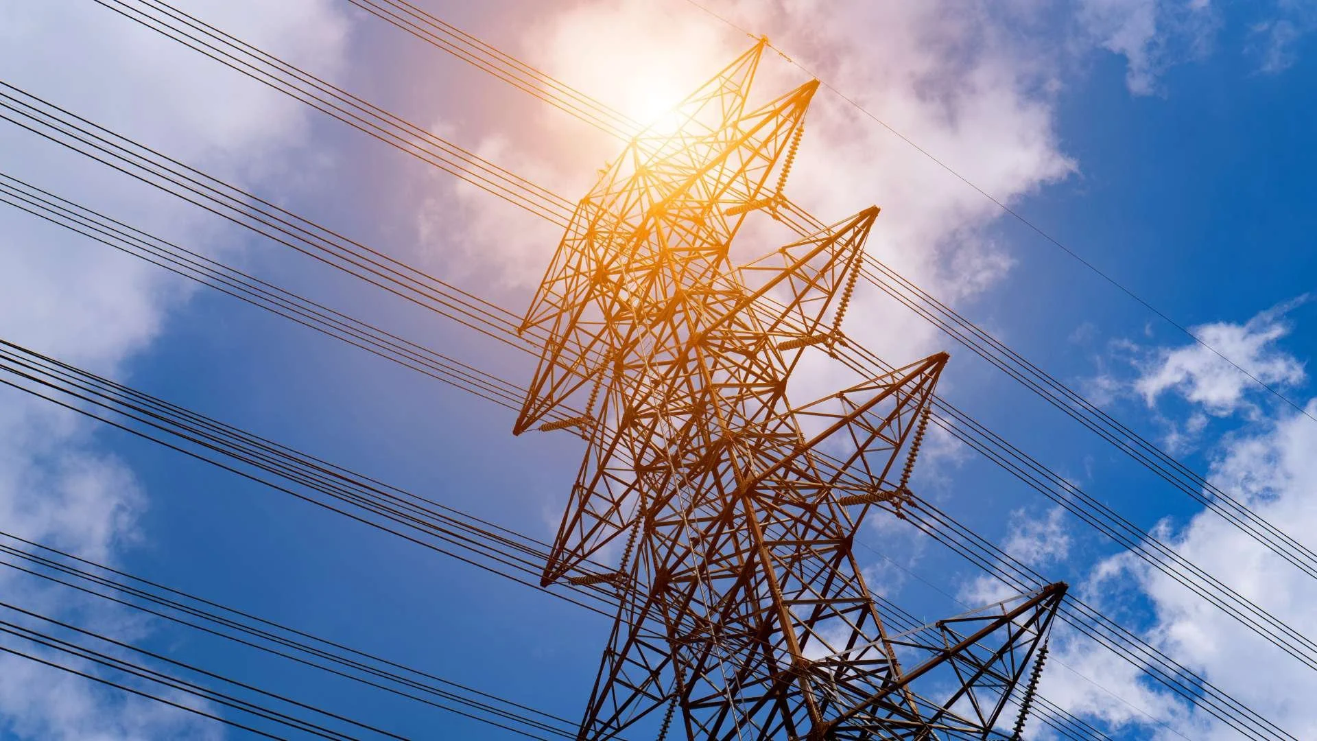 High-voltage electrical transmission tower under bright summer sunlight representing increased energy demand and system strain during warmer weather conditions
