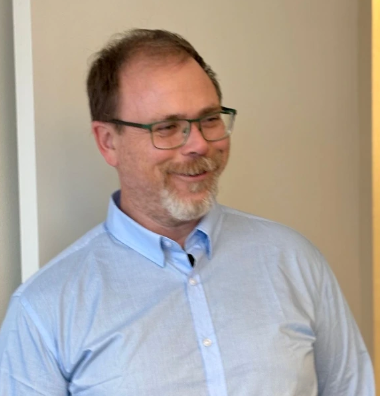 Man with glasses smiling, wearing a light blue button-up shirt, in an indoor setting.