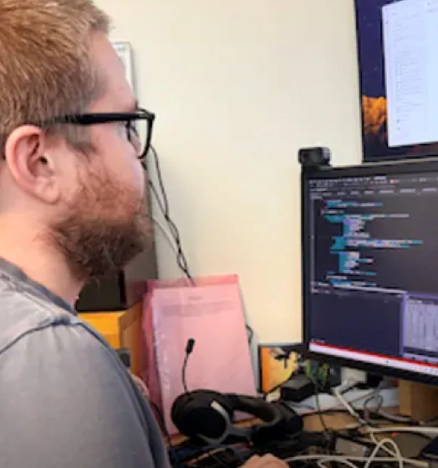A man with glasses and red hair working at a computer with dual monitors, editing code on one screen.
