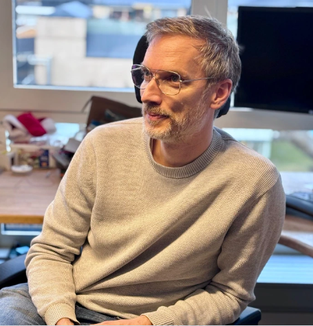 A middle-aged man with glasses and gray hair, wearing a beige sweater, sitting in an office with a desk, computer monitor, and window in the background.