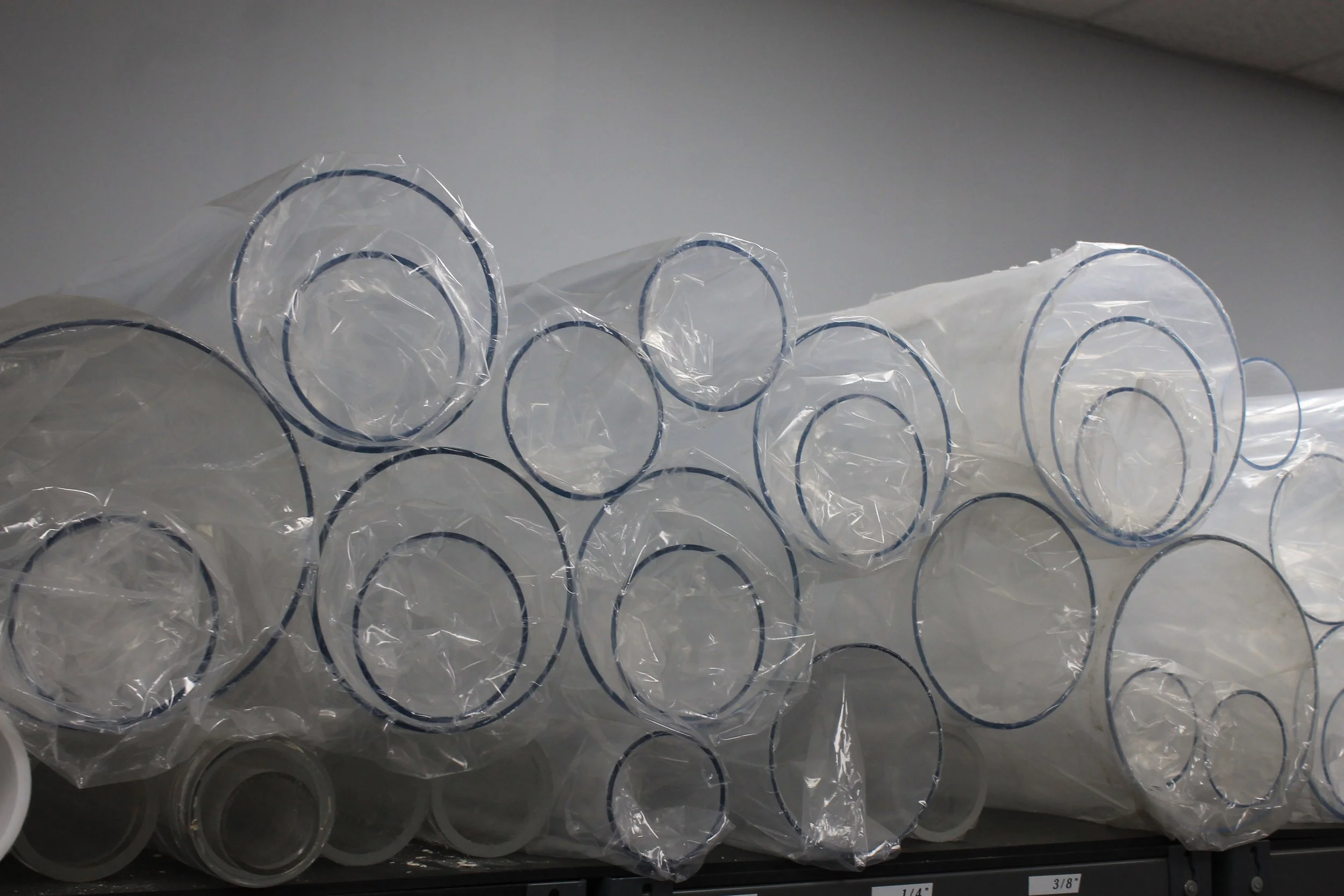 Stack of clear plastic tubes with blue circles at the ends, wrapped in plastic, stored on a shelf.