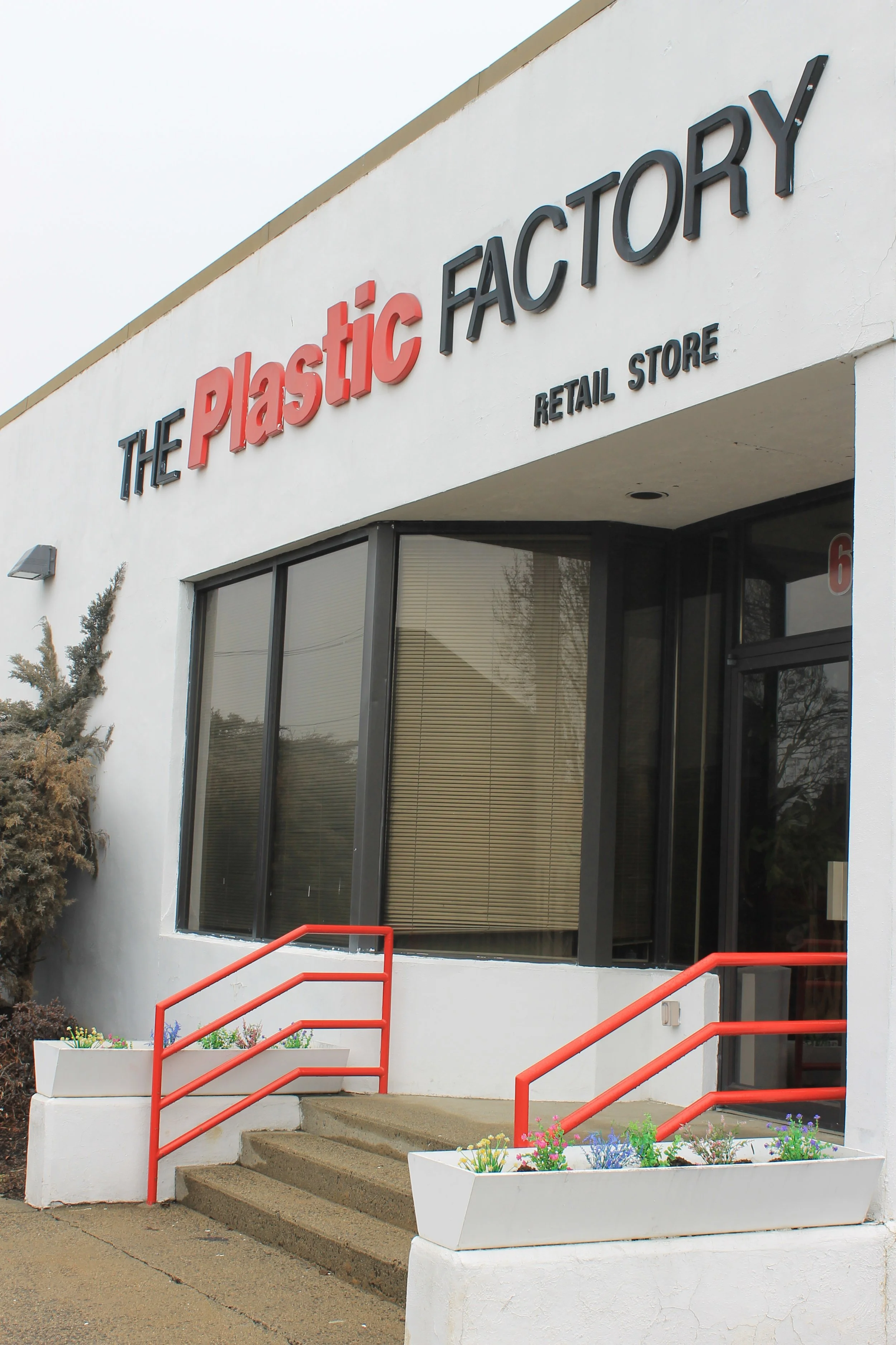 The storefront of The Plastic Factory retail store with large windows, a glass door, and red handrails on the concrete stairs leading up to the entrance, with flower planters on each side.