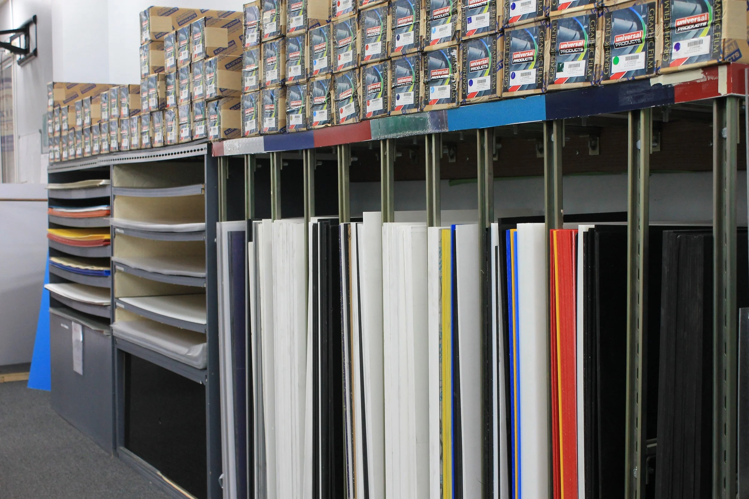Display of various stacks of vinyl and cardboard display materials in an office supply store, with top shelves of boxes of universal graphic products. Based on the stored items, it appears to be a display of signage or display boards.